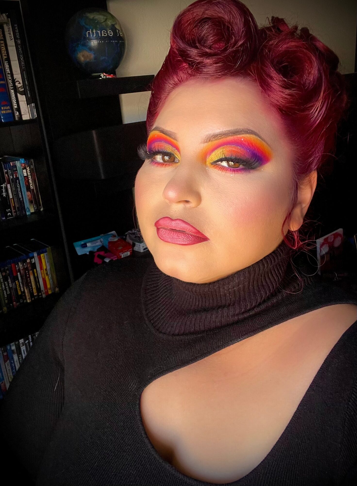 Person with vibrant rainbow makeup, styled red hair, wearing a black turtleneck with a cutout, in a room with bookshelves