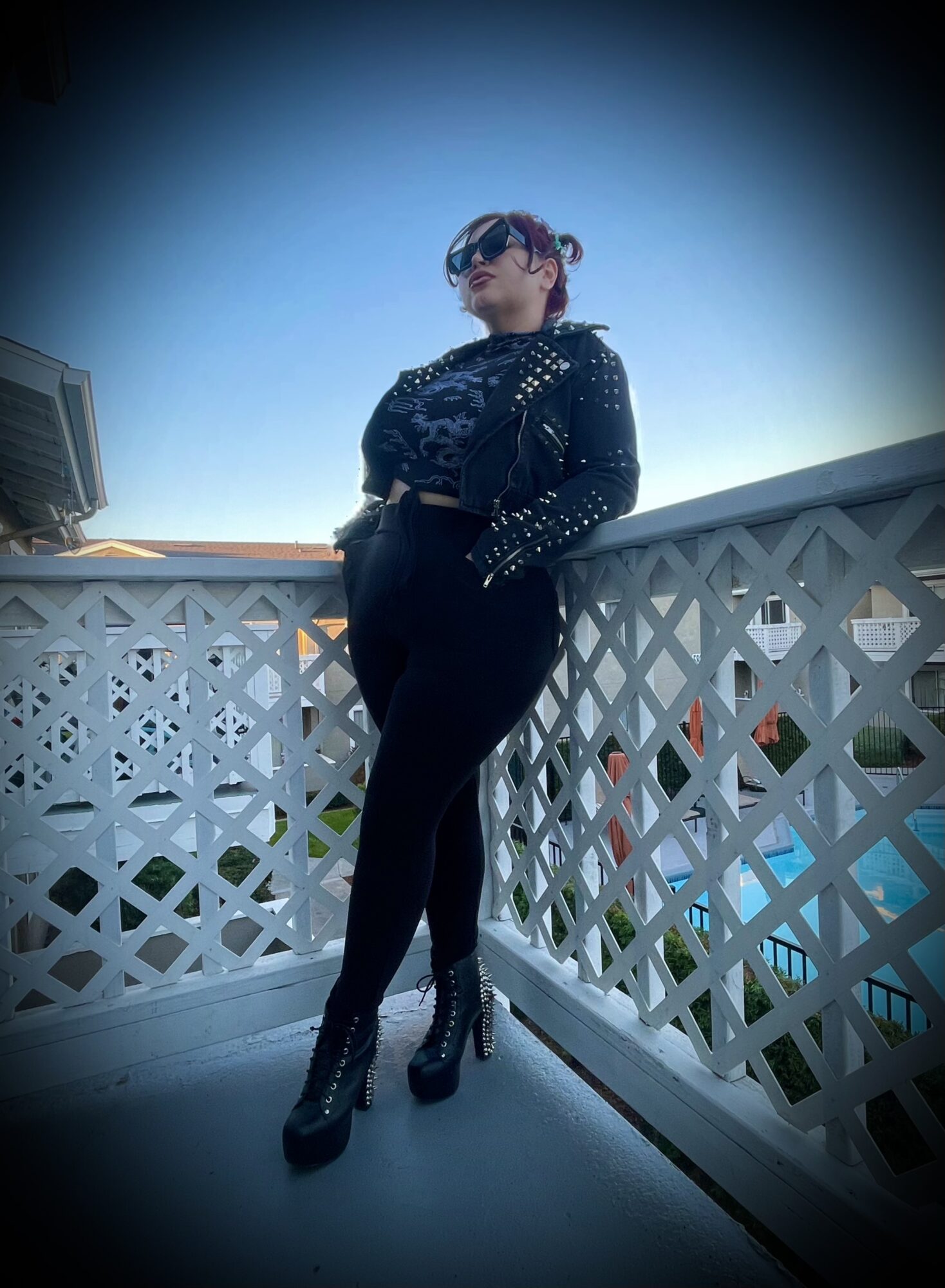 Person leaning on a white lattice railing outdoors, wearing sunglasses, a studded jacket, and high-heeled boots, with a clear blue sky.