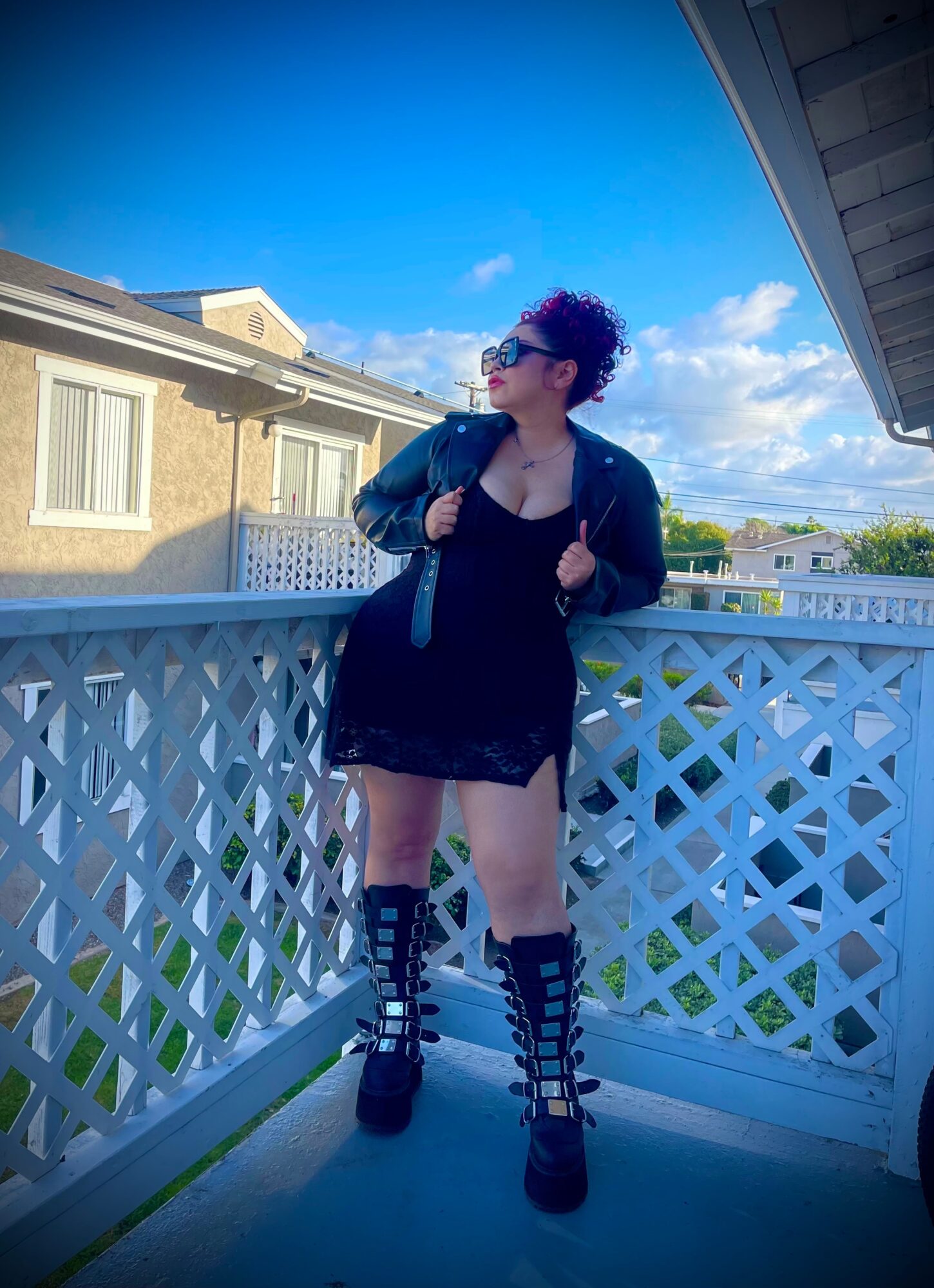 Person with curly hair and sunglasses standing on a balcony with a white lattice railing, wearing a black dress, boots, and a jacket, under a blue sky.