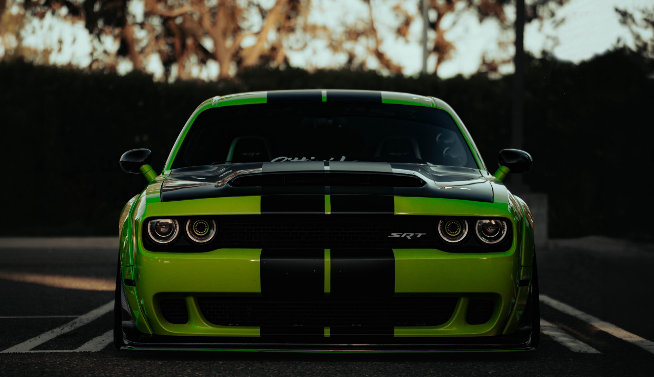 Front view of a bright green sports car with black racing stripes, parked outdoors during sunset.