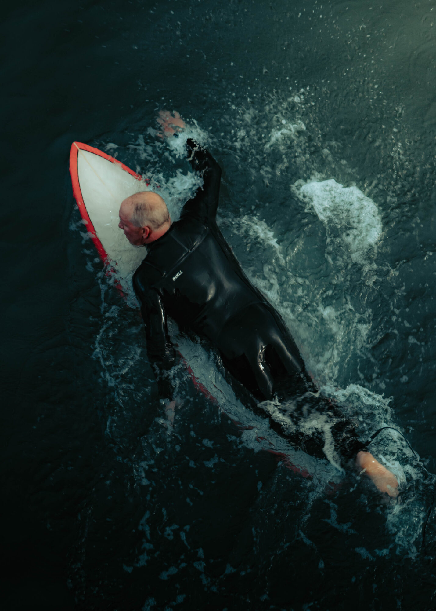 Person in a wetsuit paddling a surfboard in dark water, with splashes around them.