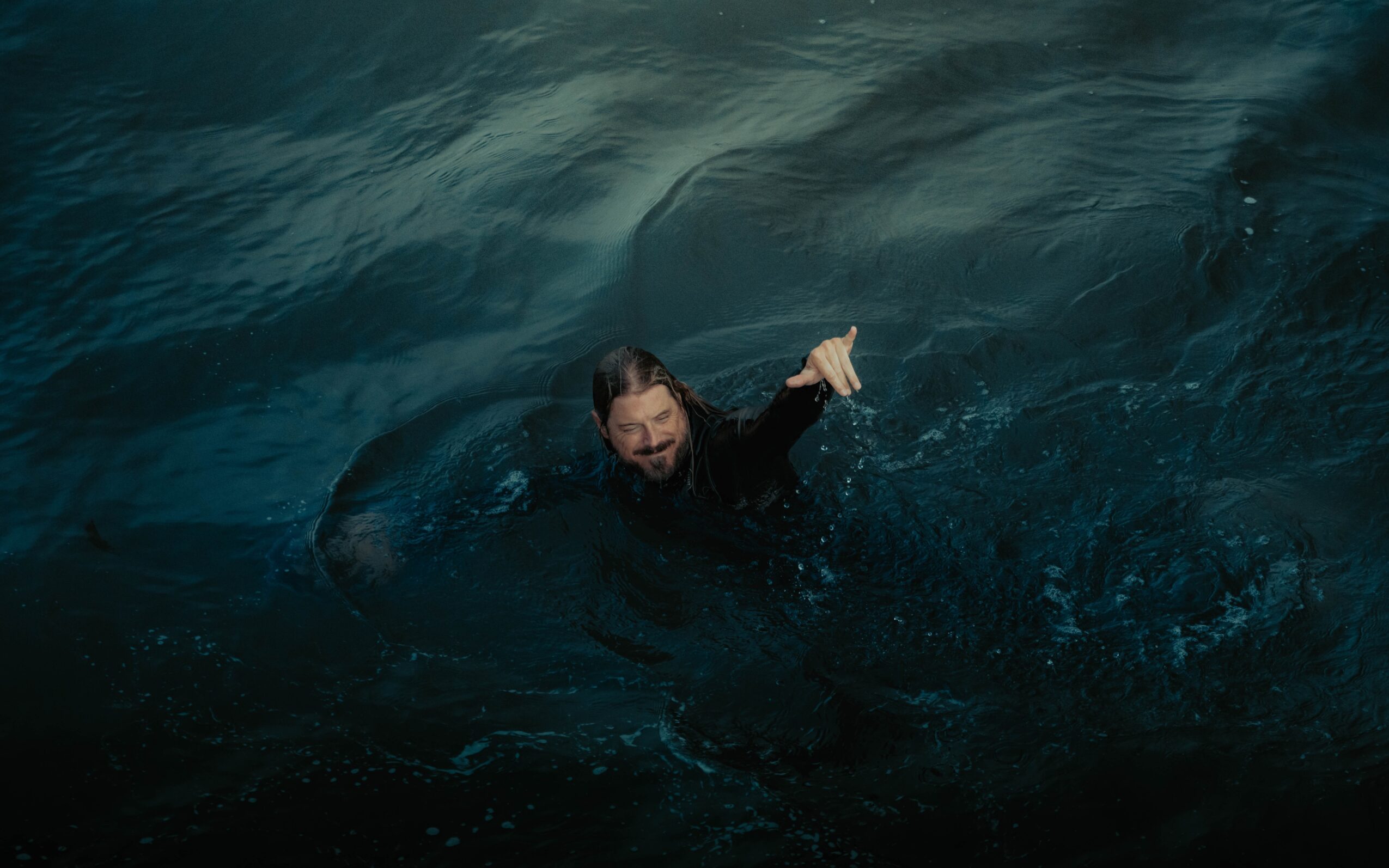 Person swimming in dark water, smiling and giving a thumbs-up, with water splashing around.