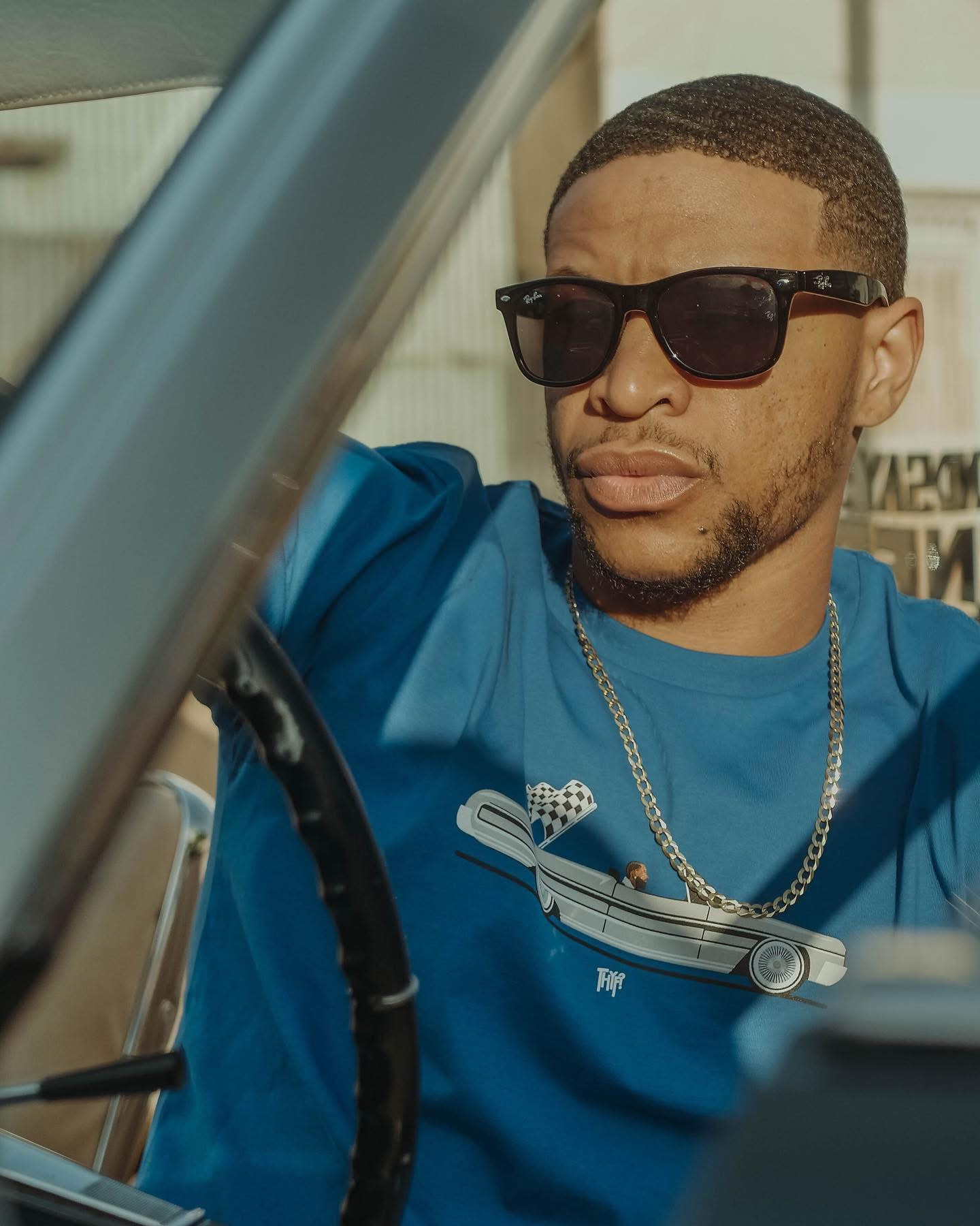 Man wearing sunglasses and a blue shirt, sitting in a vehicle, with a serious expression.