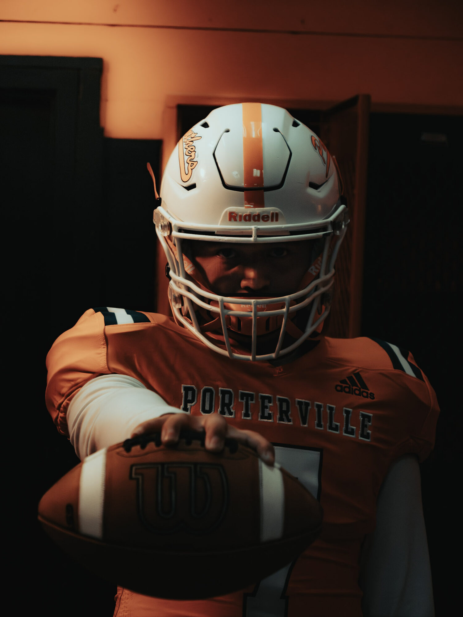 Football player in orange jersey and white helmet reaching forward with football, indoor setting, dim lighting