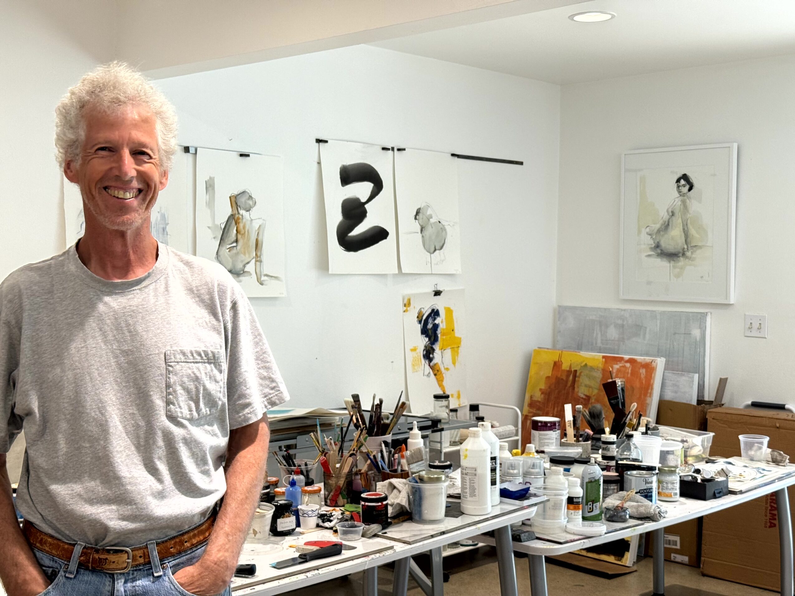 Elderly man with curly white hair smiling in art studio with paintings and art supplies on table.
