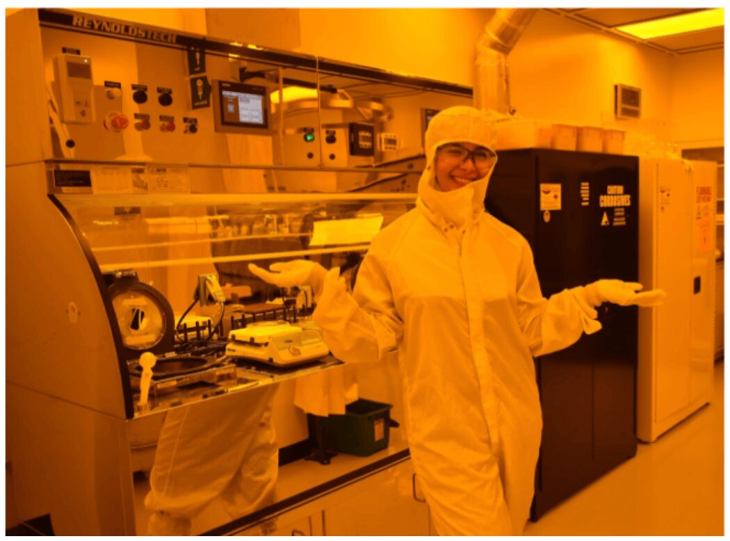 Person in protective clothing smiling in laboratory with equipment and machinery behind.