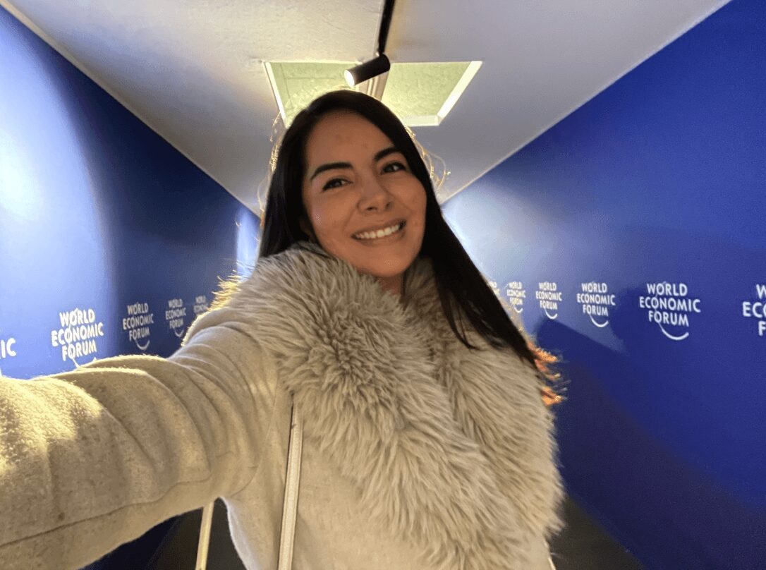 Young woman with long dark hair smiling, wearing a light-colored fur coat, taking a selfie in a blue-walled corridor.