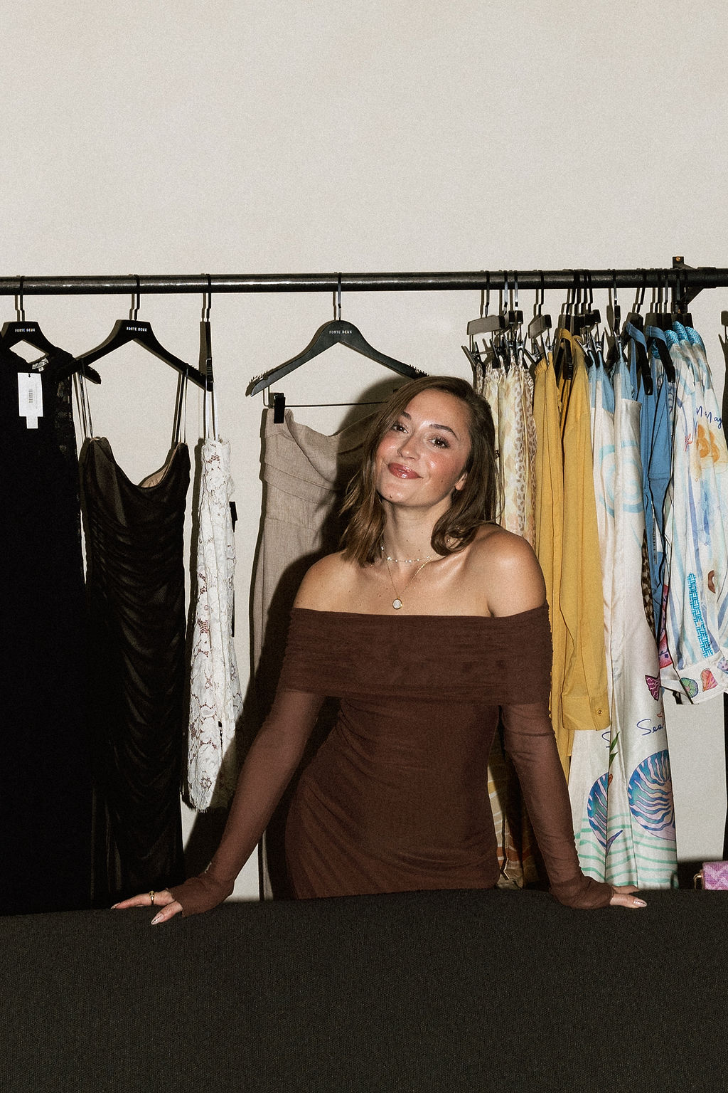Young woman with shoulder-length hair in a brown dress standing in front of a clothing rack with various dresses and garments.