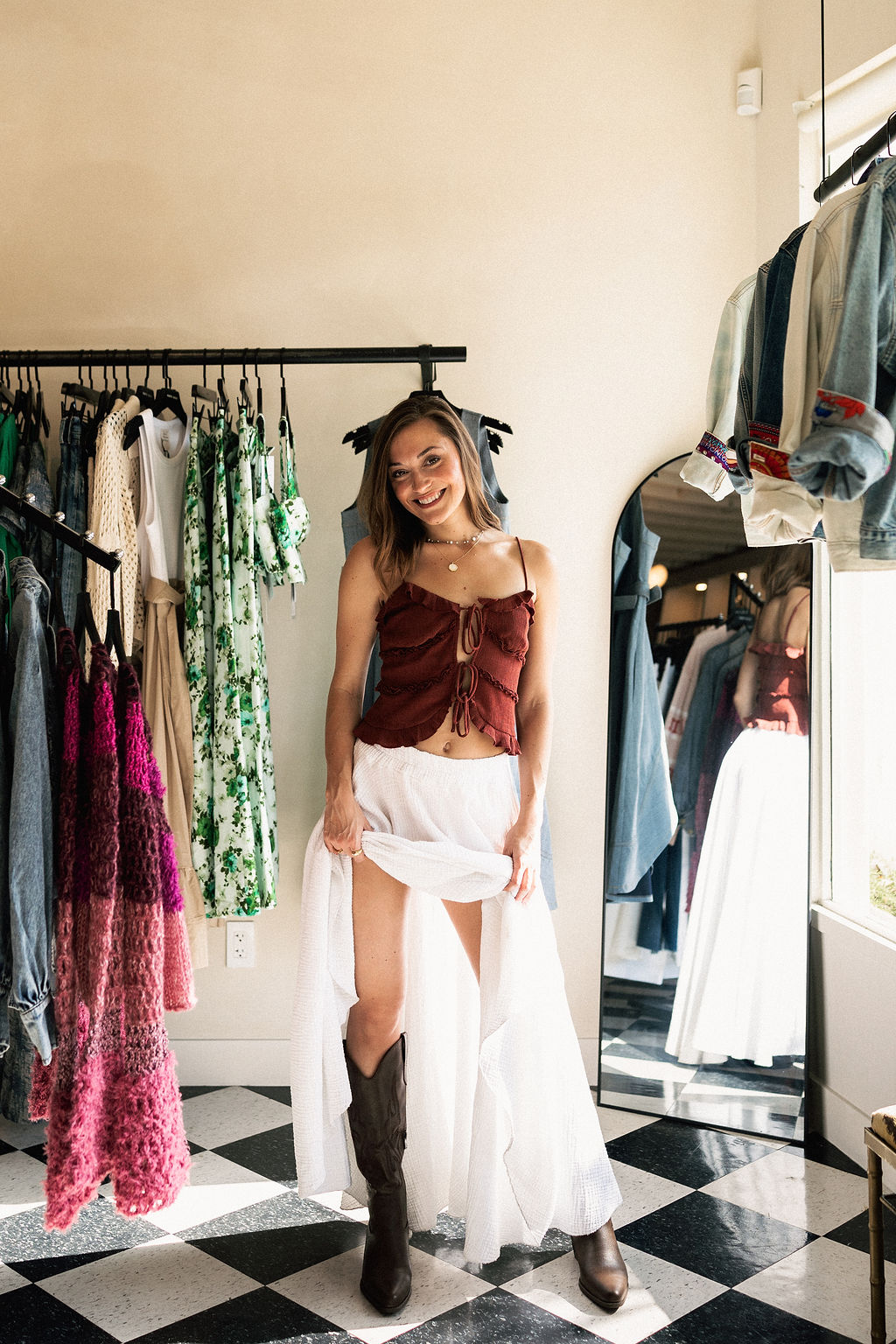 Woman in a boutique trying on a white skirt and brown top, surrounded by clothing racks and a mirror.