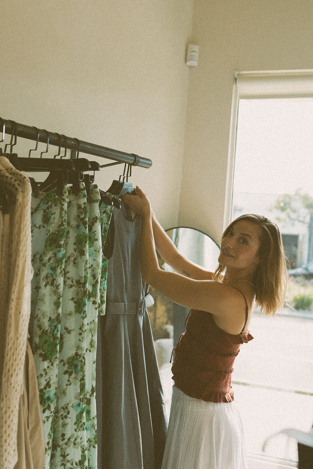 Young woman with shoulder-length hair wearing a sleeveless top and white skirt, shopping for dresses in a store, holding a dress on a hanger.