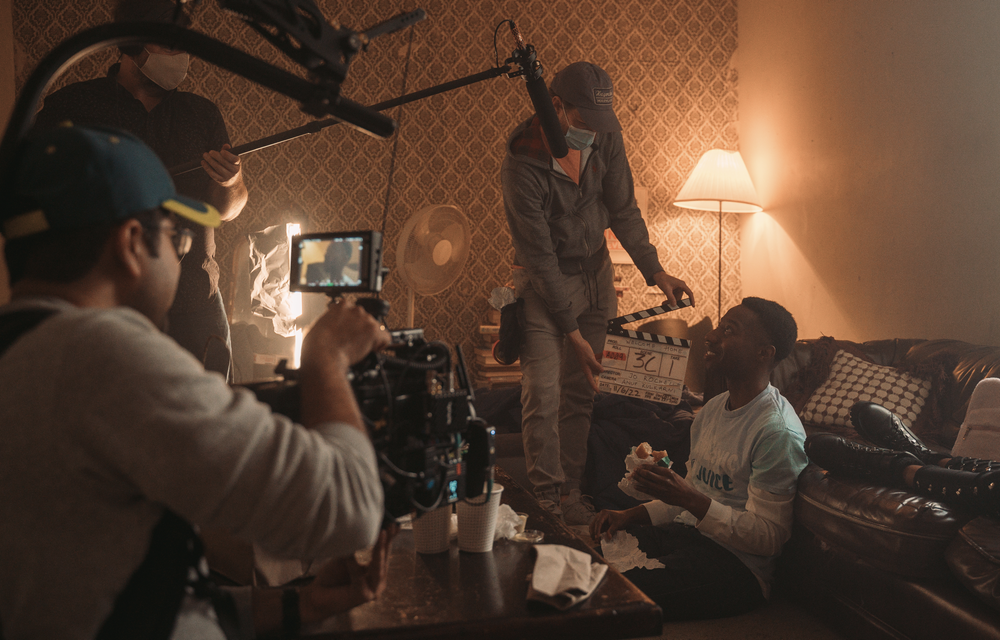 Filming crew setting up scene with two people in a dimly lit room, one sitting and one standing, with equipment and a lamp.