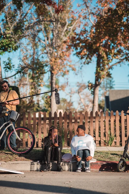 Two children sit on a sidewalk, one with a backpack, in front of a wooden fence with trees and blue sky behind.