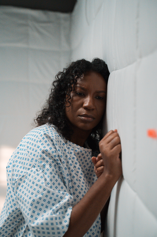 Woman with curly hair leaning against a padded wall, wearing a hospital gown, looking at the camera.