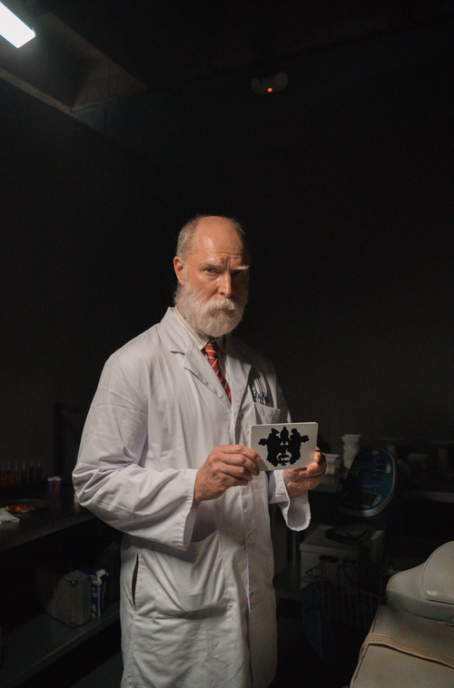 Older man with glasses and a beard in a white lab coat holding a small object, standing in a dark room.