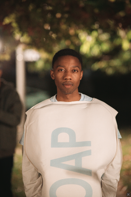 Young person with short hair wearing a white jacket with large light blue letters, standing outdoors at night.