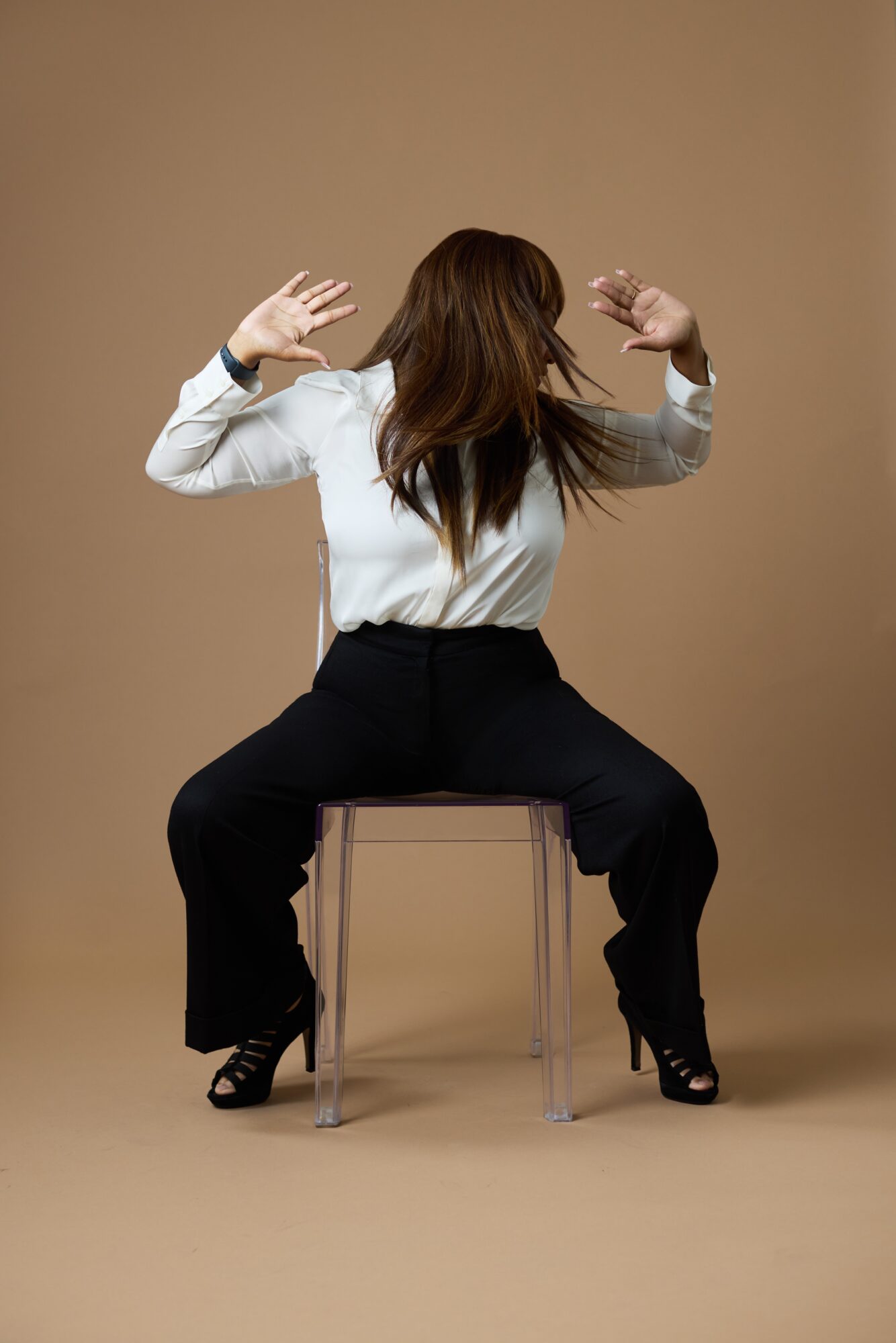 Person with long hair sitting on a transparent chair, facing away, with arms raised and hair covering face.