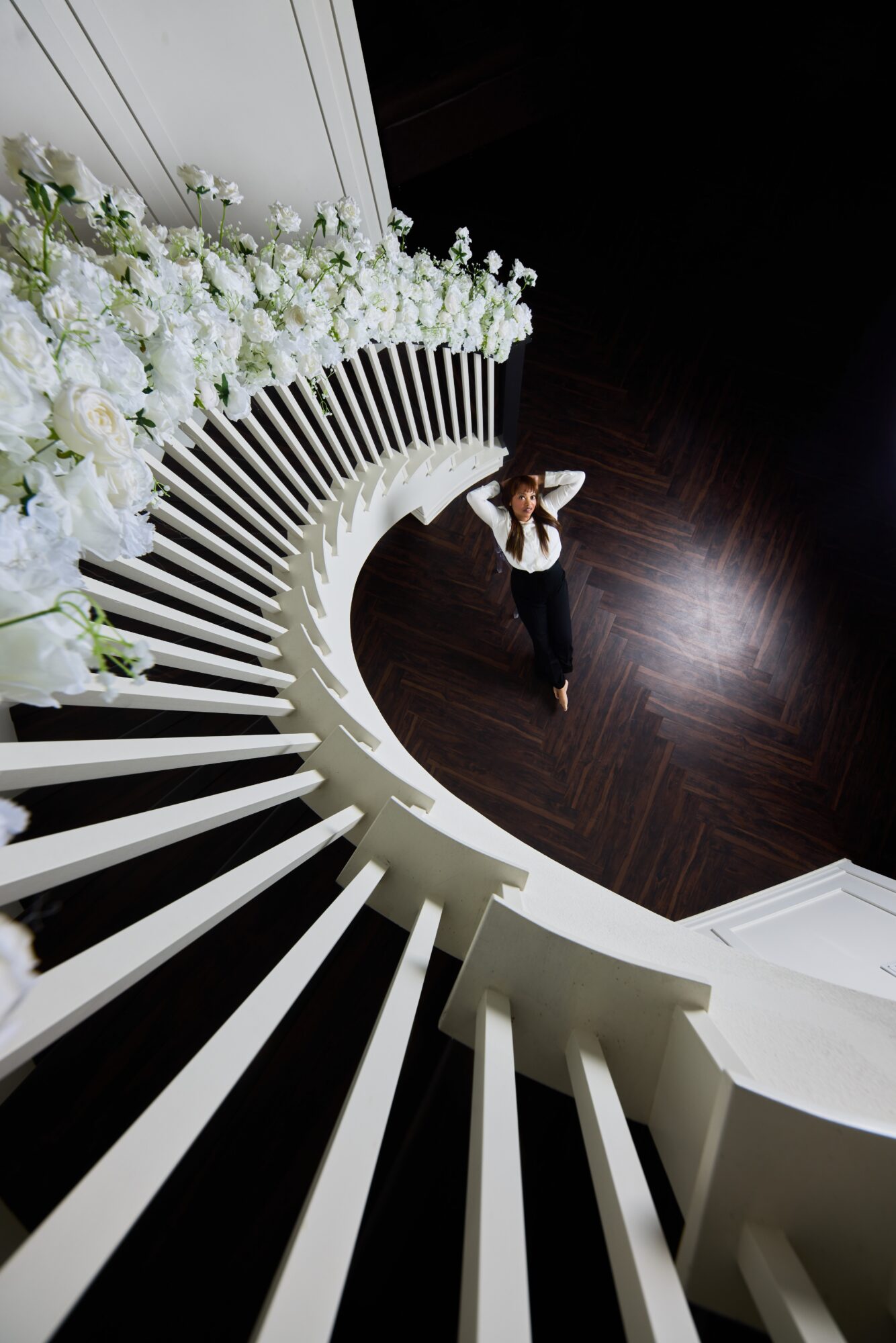 Person standing at the bottom of a curved staircase with white railings and flowers, viewed from above.