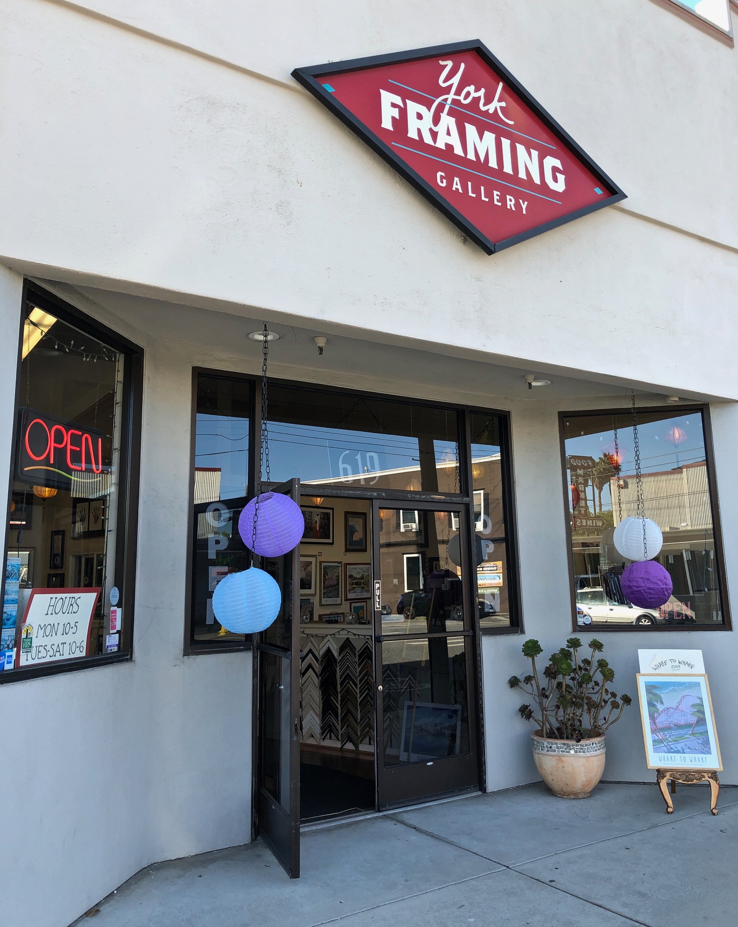 Gallery entrance with open door, purple and blue balloons, potted plant, and an 'Open' sign in window.