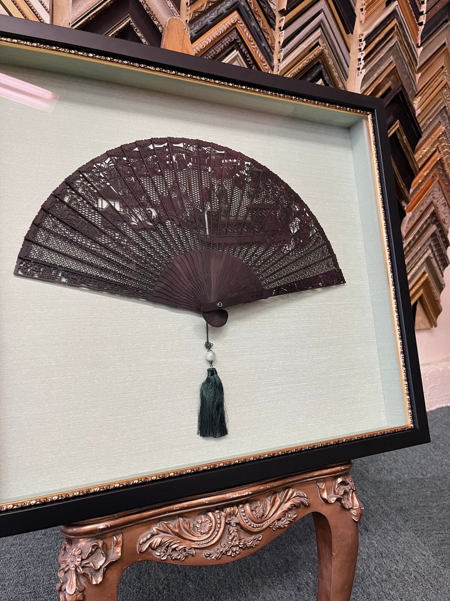 Decorative black hand fan with tassel displayed in a frame, against a background of wooden picture frames.