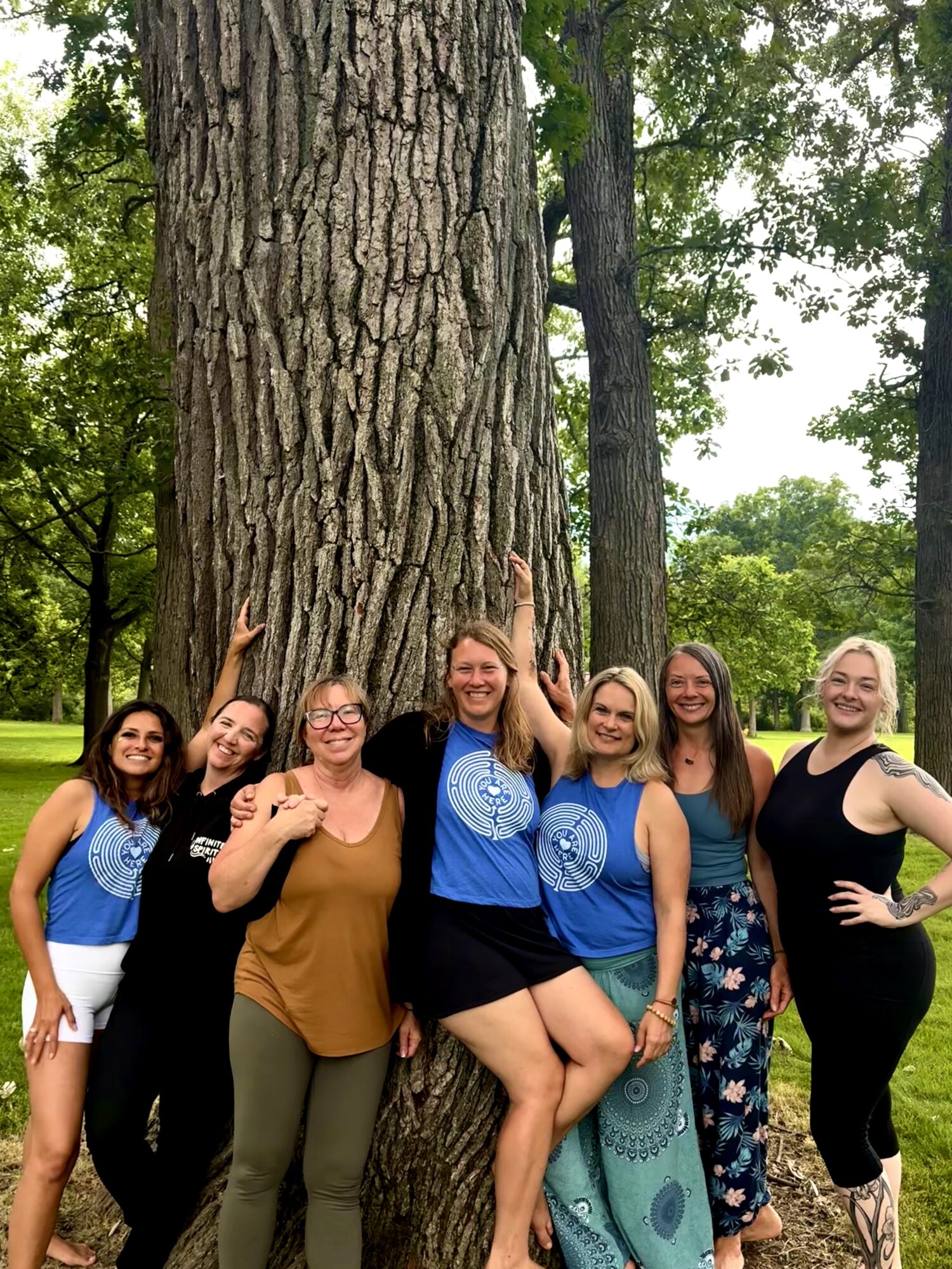 Group of six women standing outdoors near a large tree, smiling and posing for a photo.