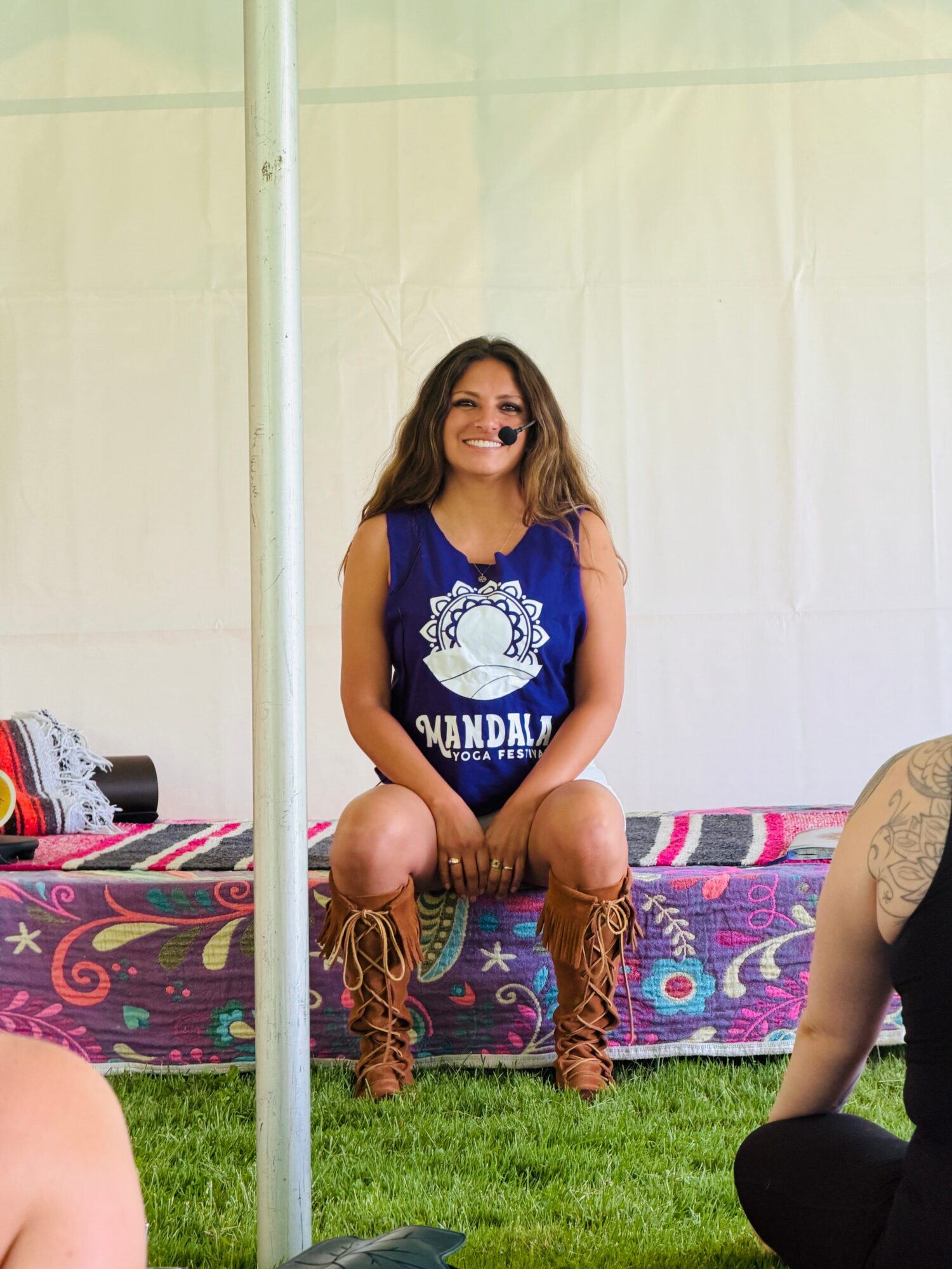 Woman sitting on a colorful bed outdoors, smiling, wearing a blue sleeveless top and brown boots, with a grassy background.