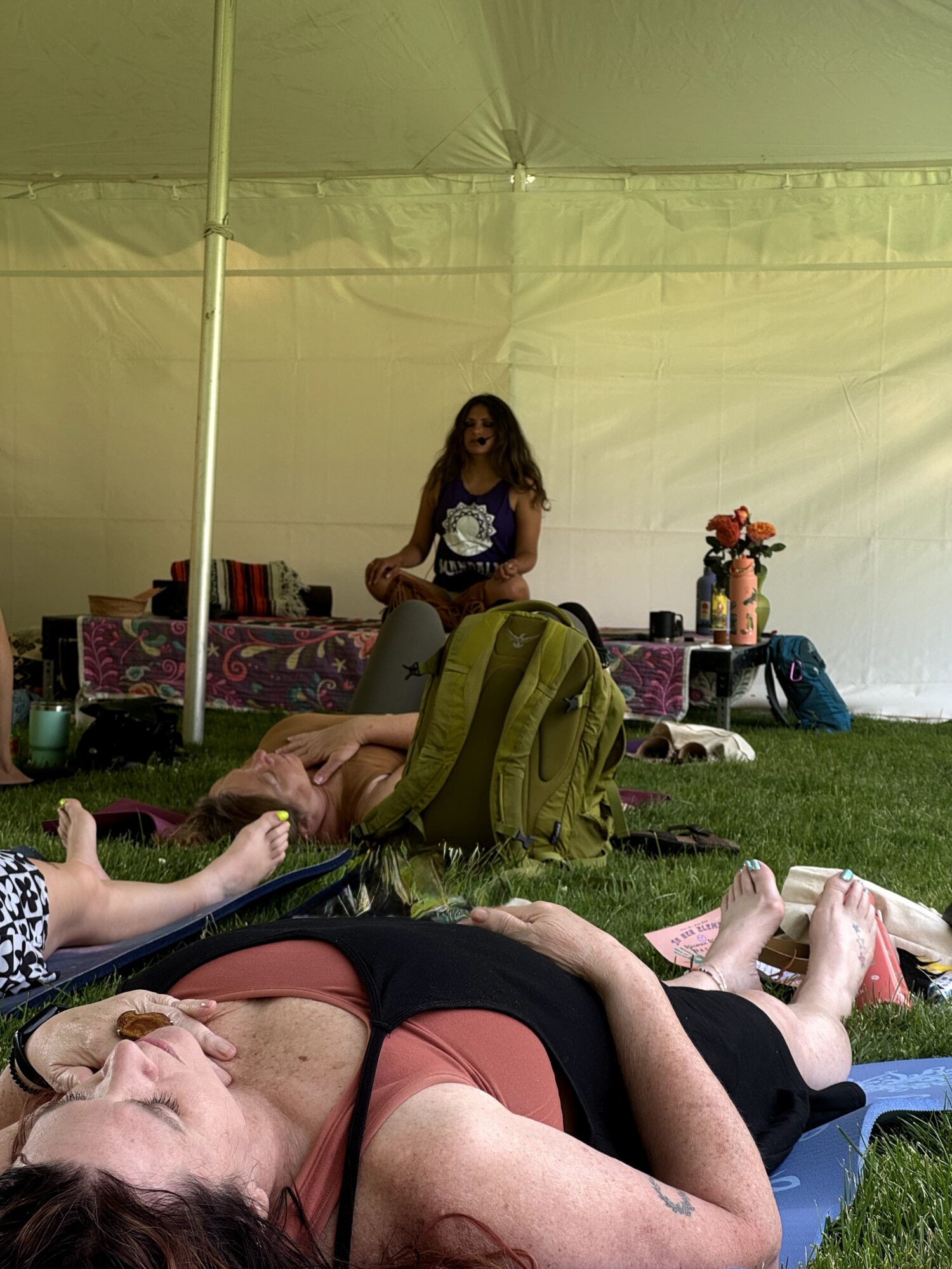 Group of people relaxing on mats inside a tent, with a woman standing in the background near a table with flowers.