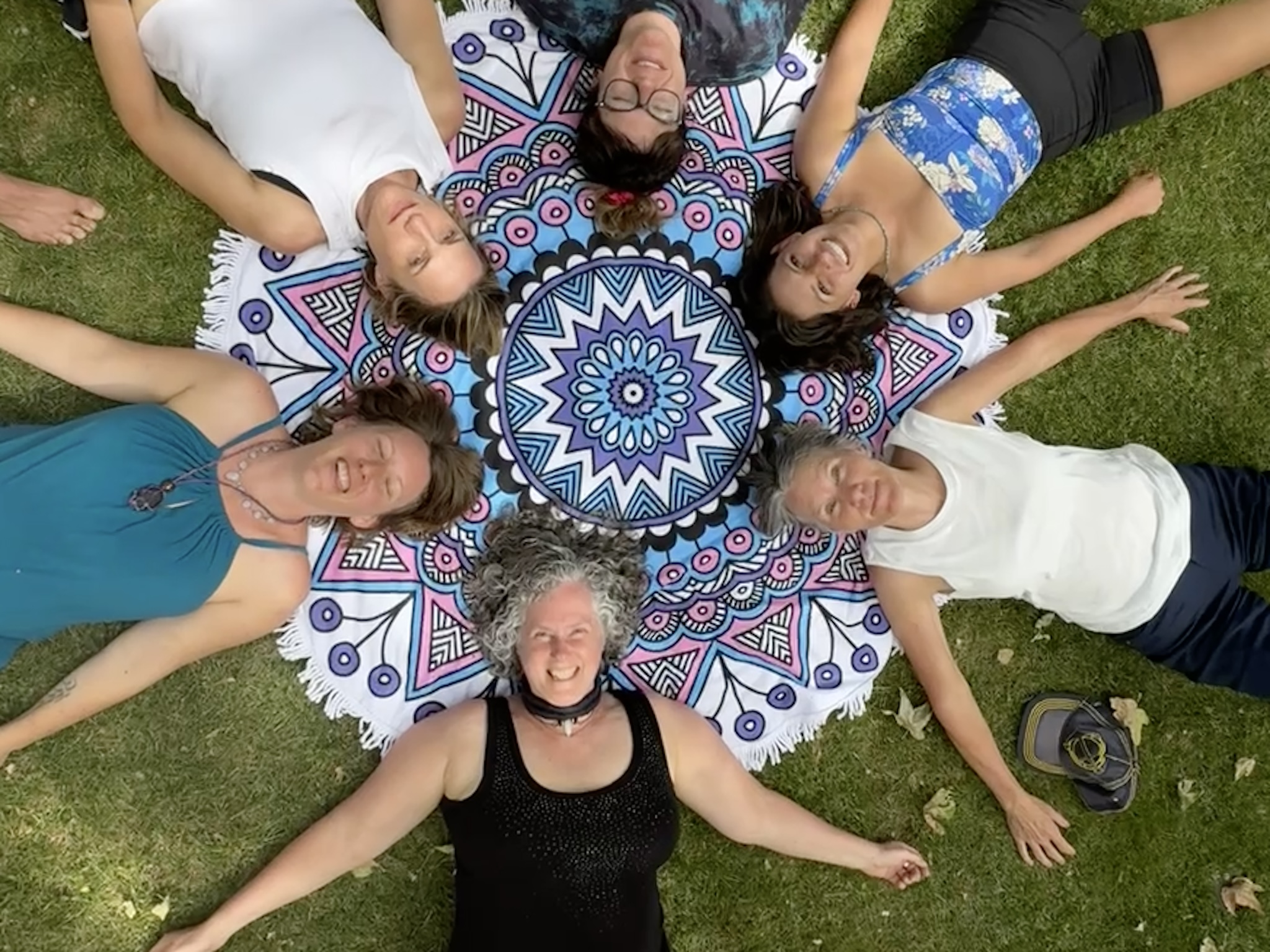 Six women lying in a circle on grass around a colorful mandala-patterned cloth, smiling and looking up at the camera.