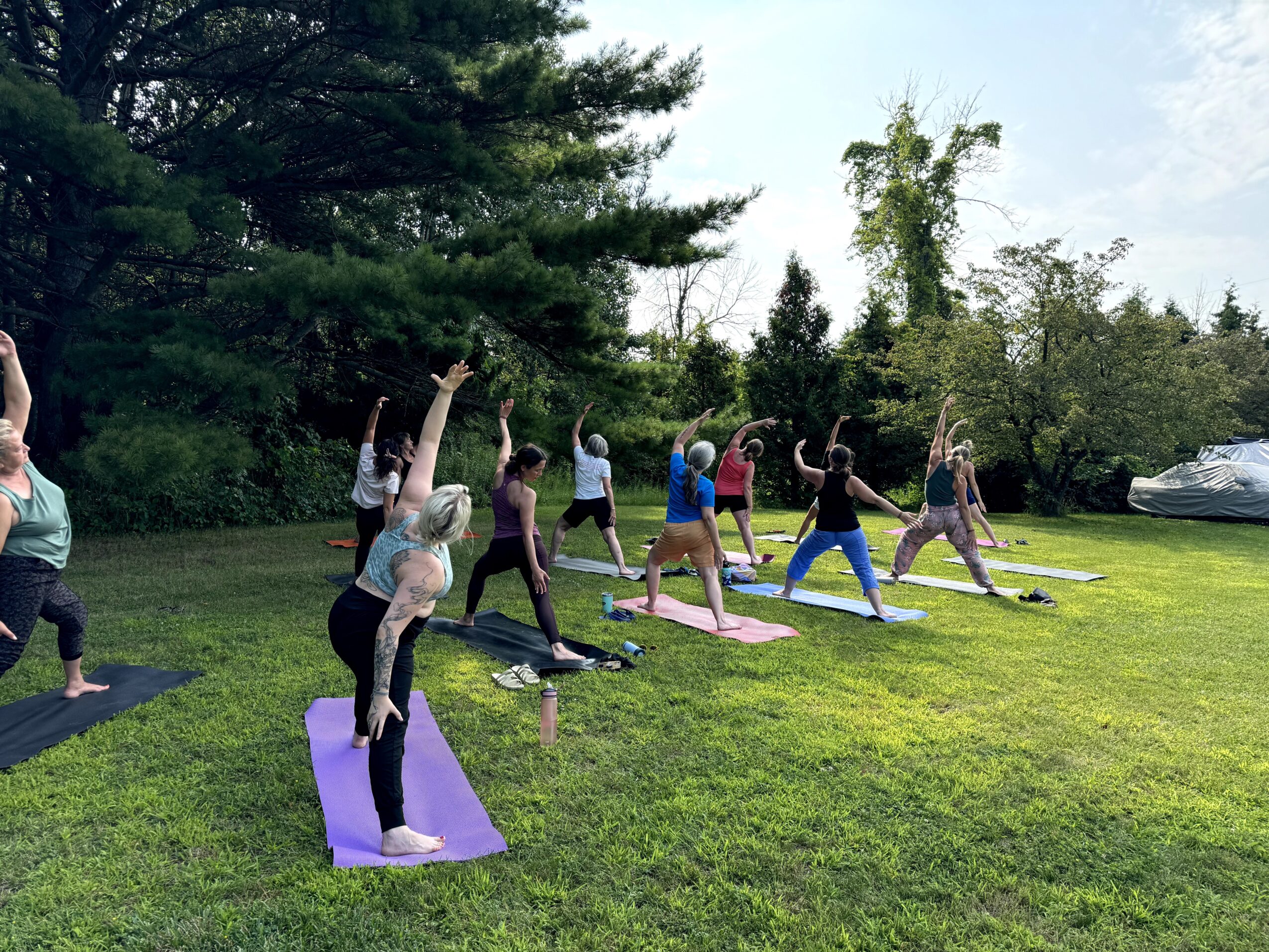Group of people practicing yoga outdoors on grass with trees in background, some on mats, in a sunny setting.