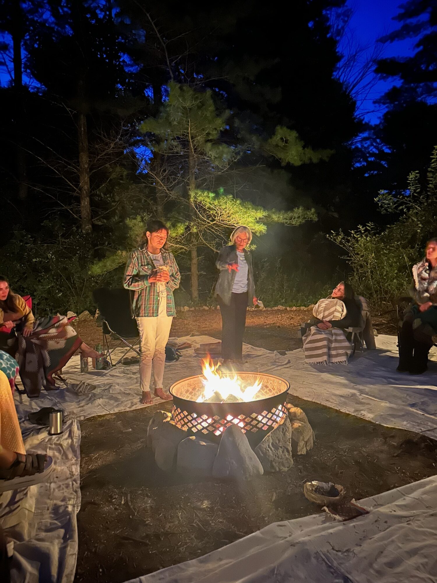 People gathered around a campfire outdoors at night, with trees in the background and some sitting on the ground.