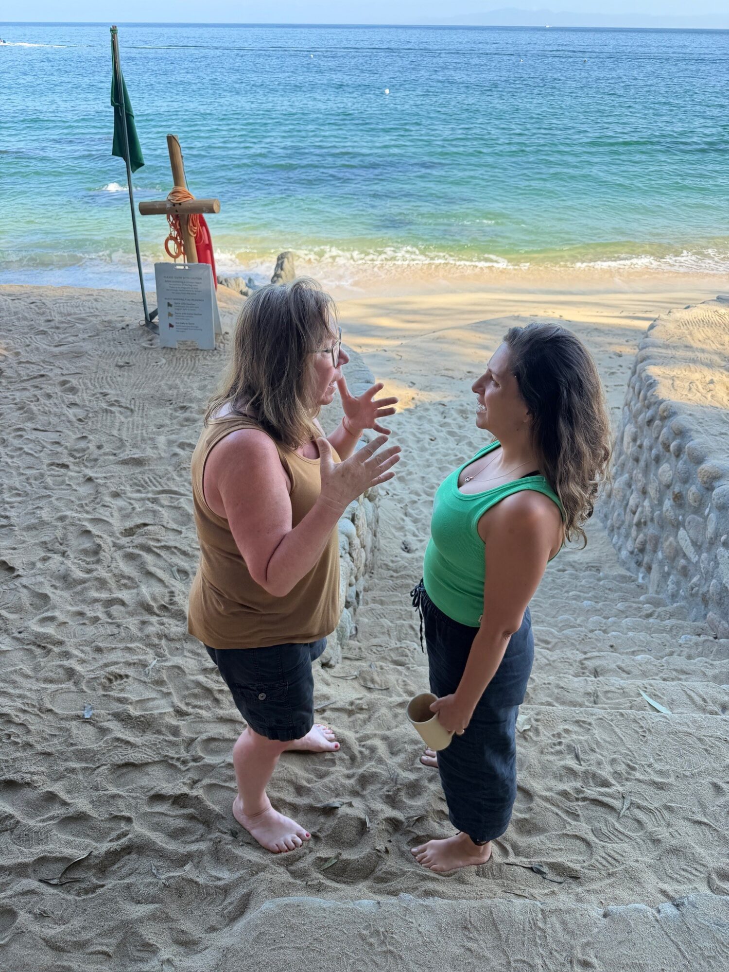 Two women stand on a sandy beach near the water, talking and gesturing, with a flag and lifeguard station in the background.