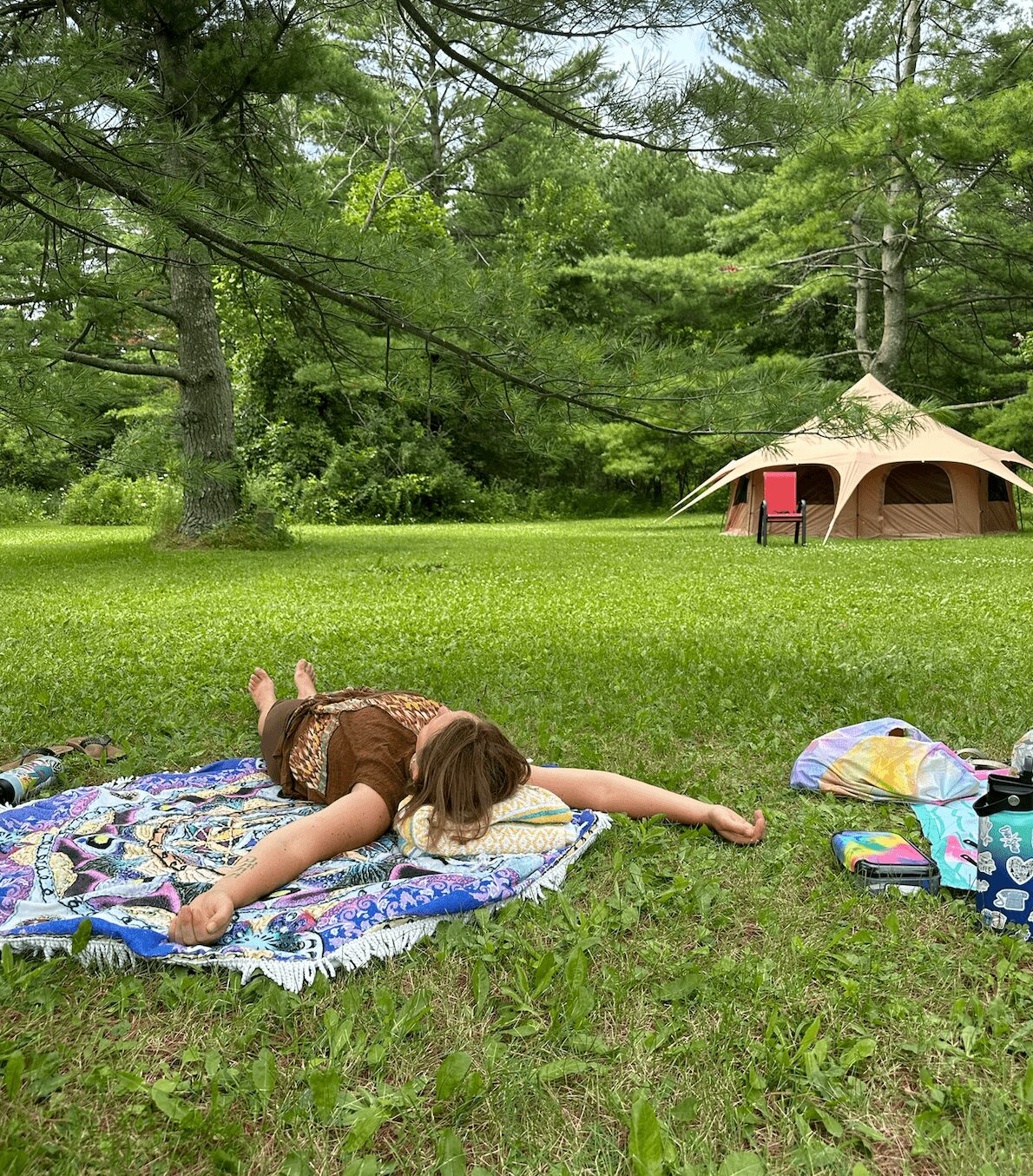 Child lying face down on a colorful blanket in a grassy area with trees and a tent in the background.
