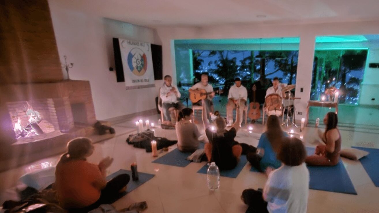 Group of people sitting on mats and chairs in a room with large windows, some playing guitars, others listening, candles on the floor.