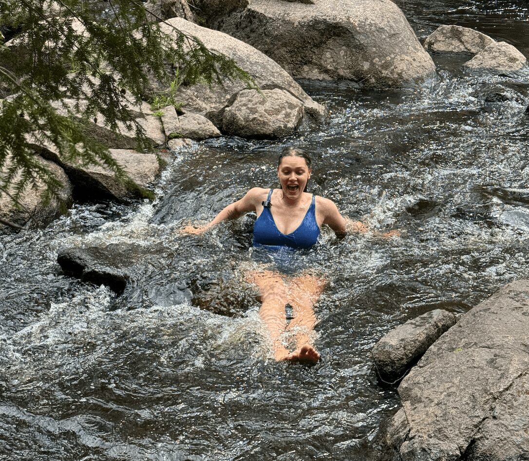 Woman in a blue swimsuit sitting in a rocky river with water flowing around her, smiling, surrounded by rocks and trees.