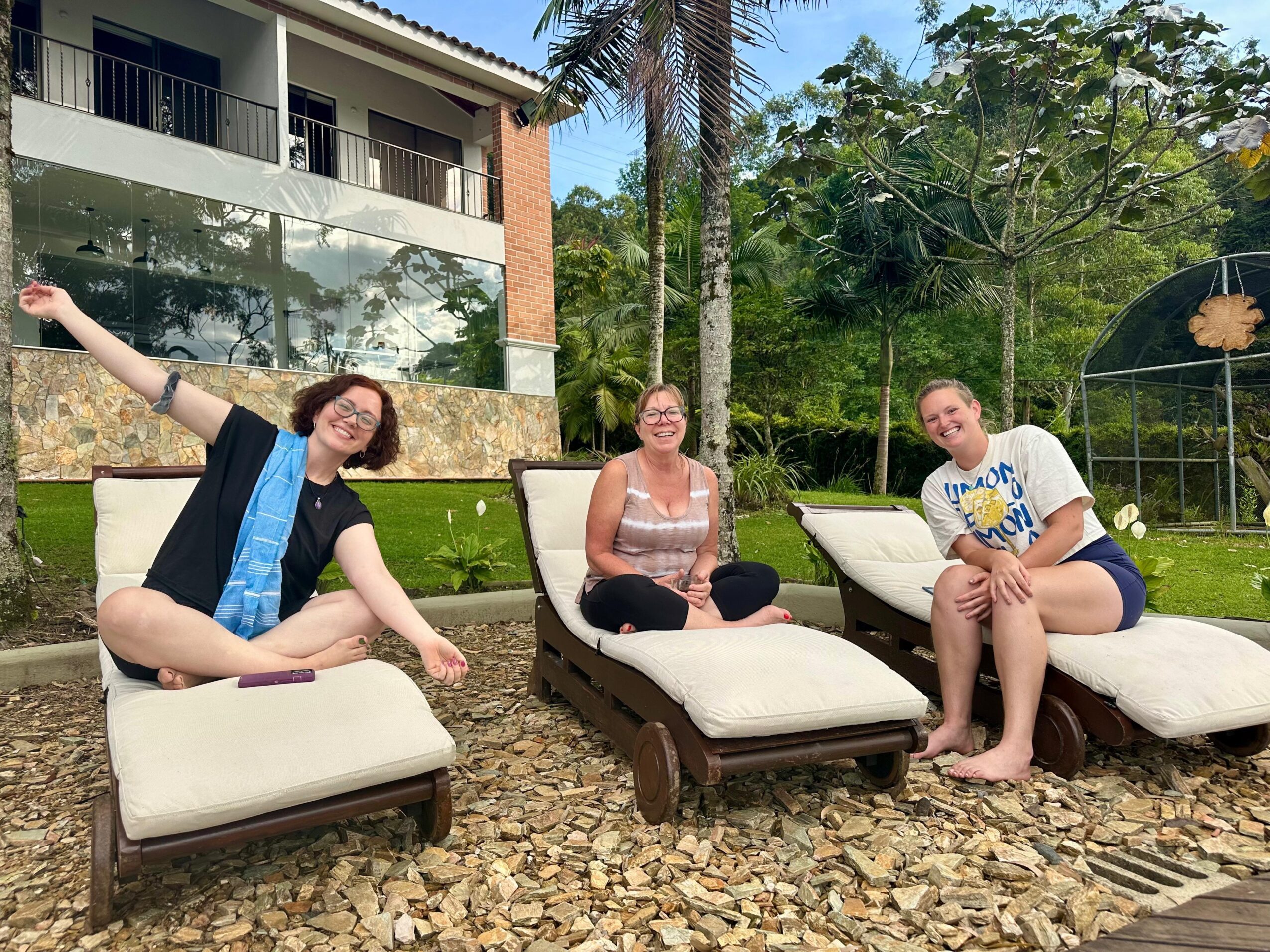Three people sitting on lounge chairs outdoors, smiling, with trees and a building in the background.