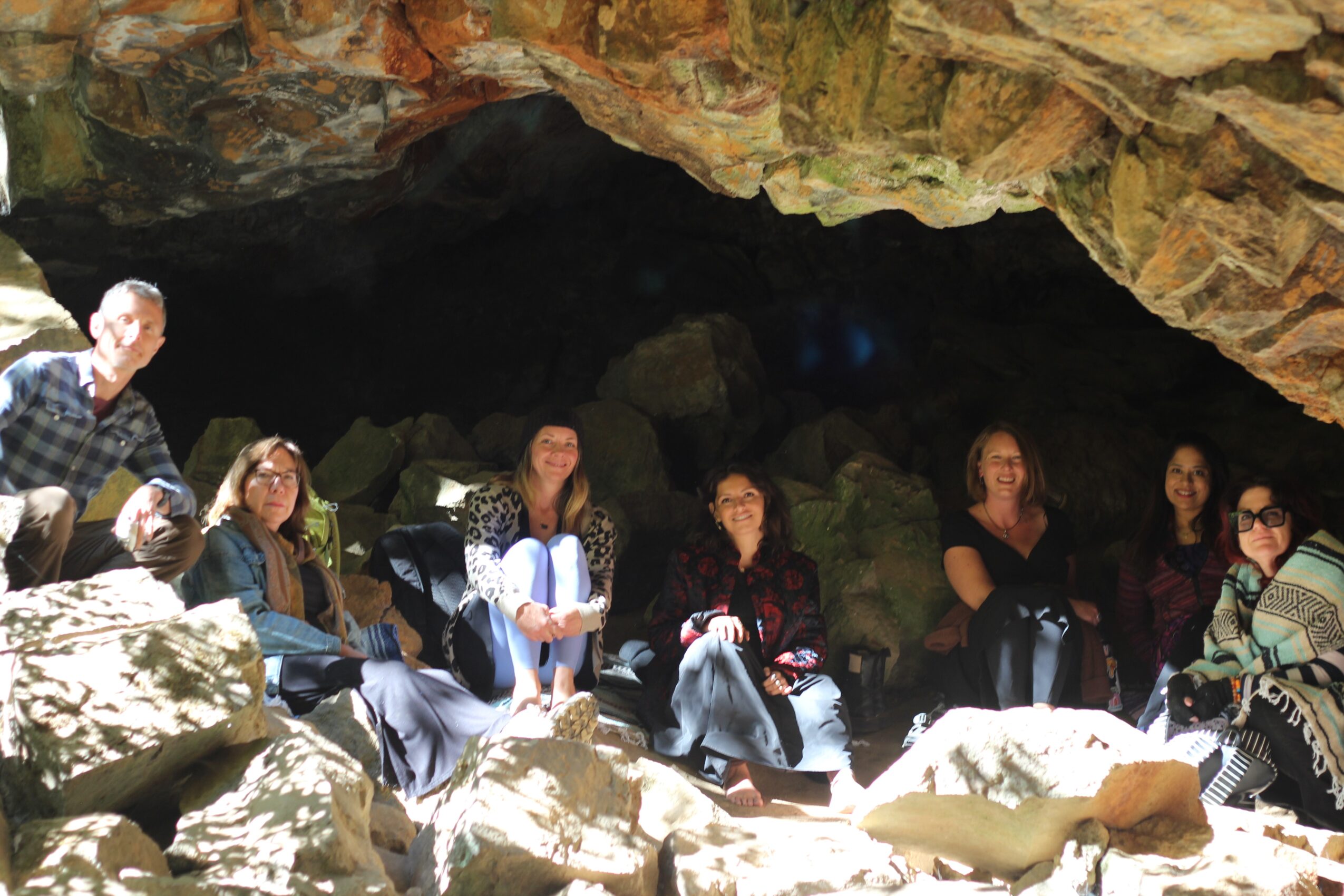Group of people sitting inside a cave with rocky walls and large rocks in front, some smiling and looking at the camera.
