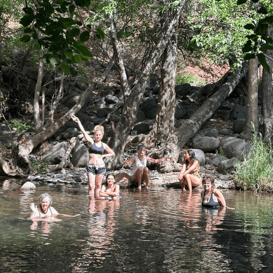 Five children playing in a shallow river surrounded by trees and rocks, some standing and some swimming.