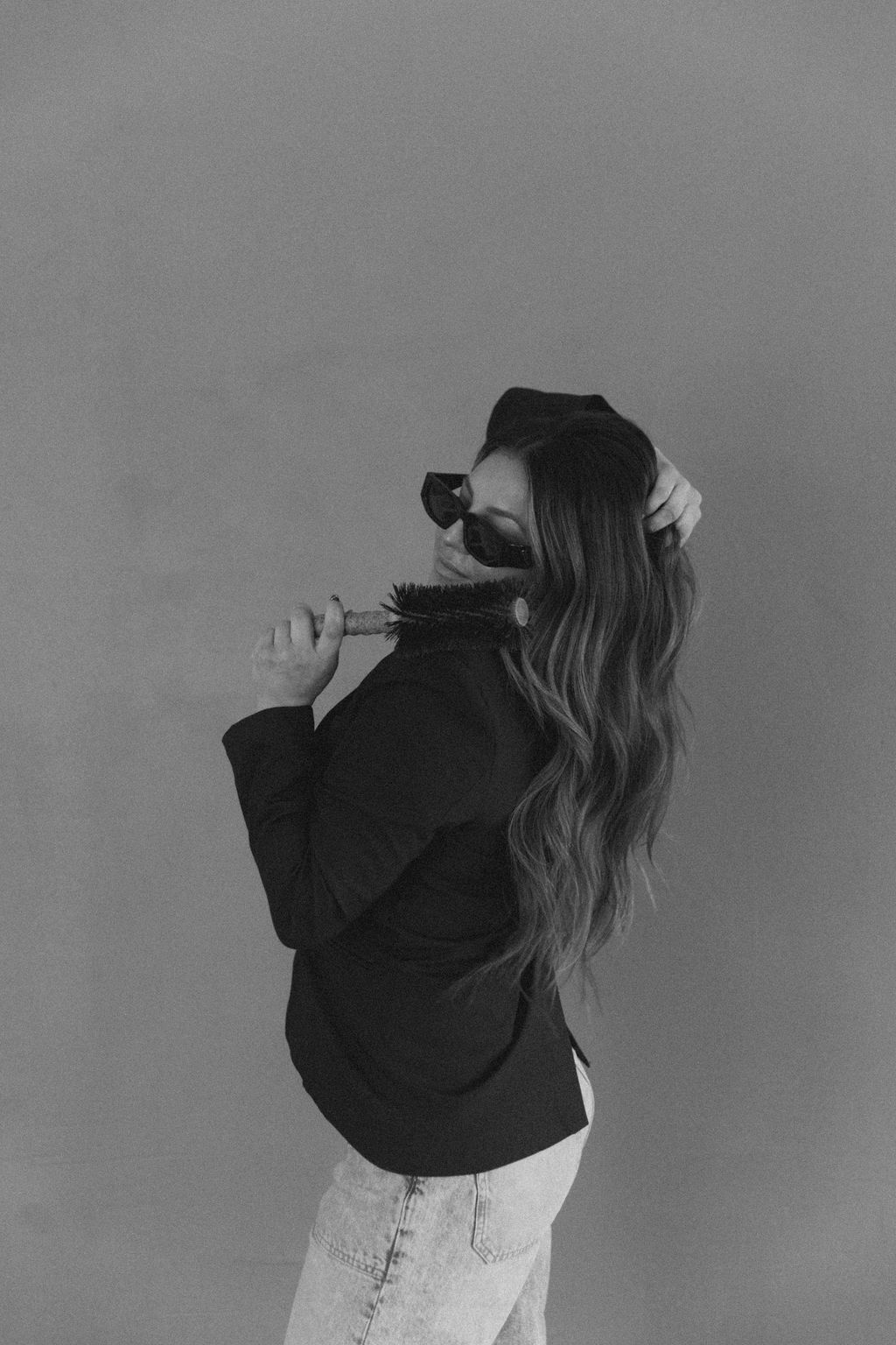 Woman with long hair wearing sunglasses and a jacket, holding a pair of glasses, standing sideways against a plain background.