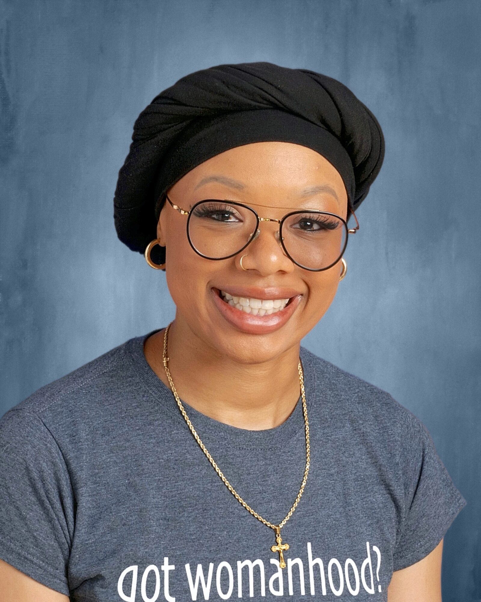 Smiling woman wearing glasses, a black headwrap, a gray T-shirt, and a gold necklace against a blue background.