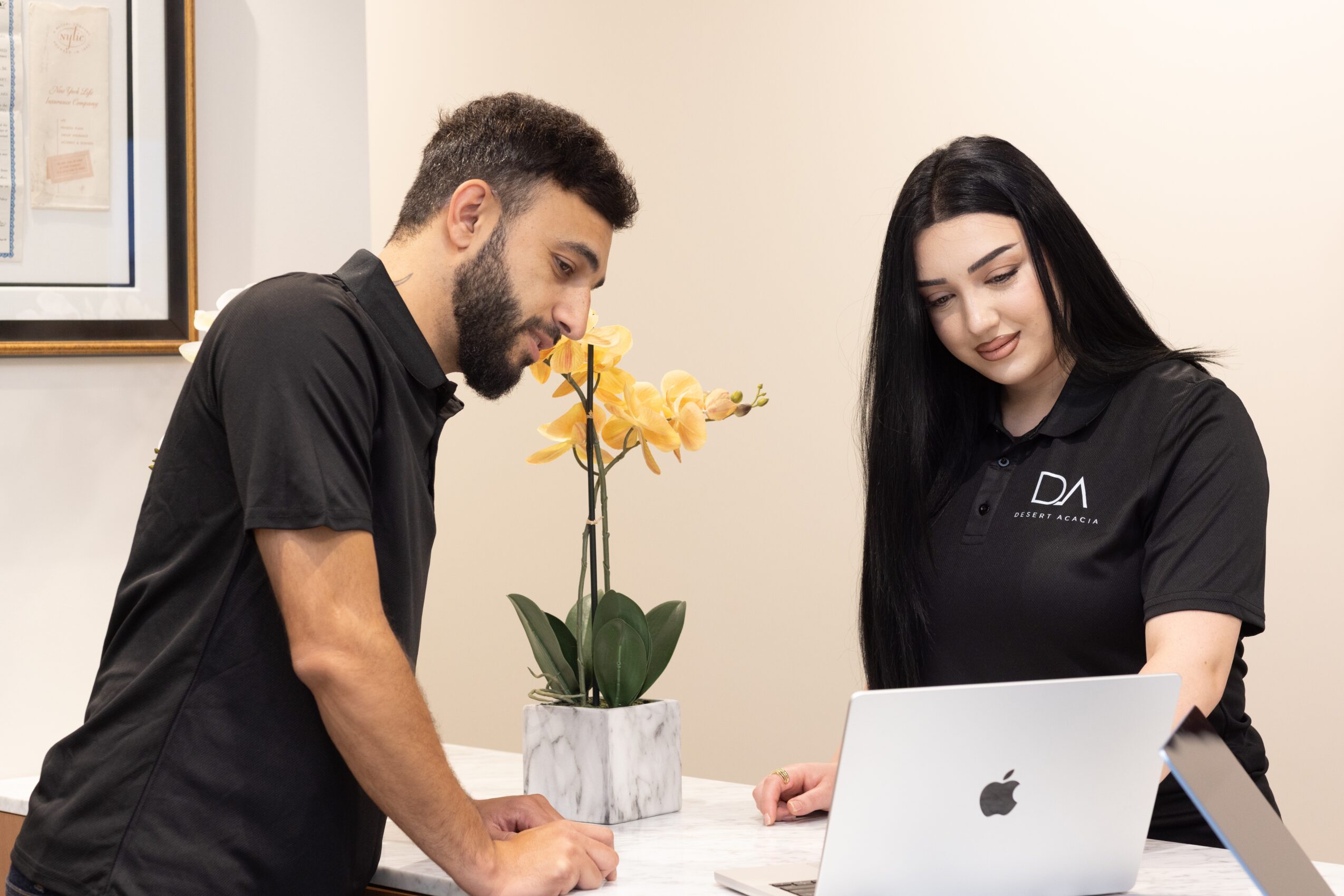 A man and woman look at a laptop screen in an office with a potted yellow flower between them.