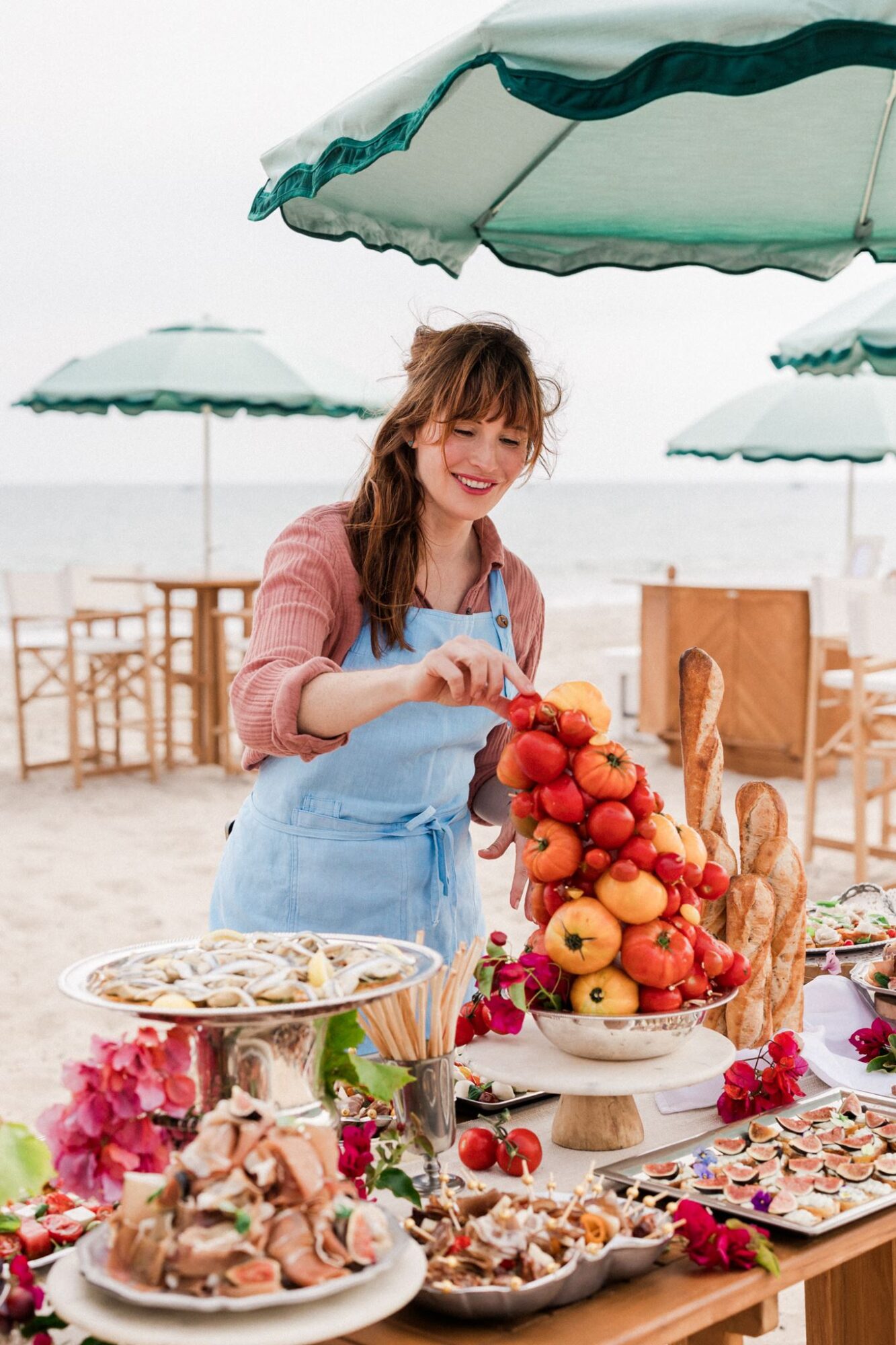 Woman arranging apples and flowers at outdoor market table on beach, with umbrellas and chairs in background.