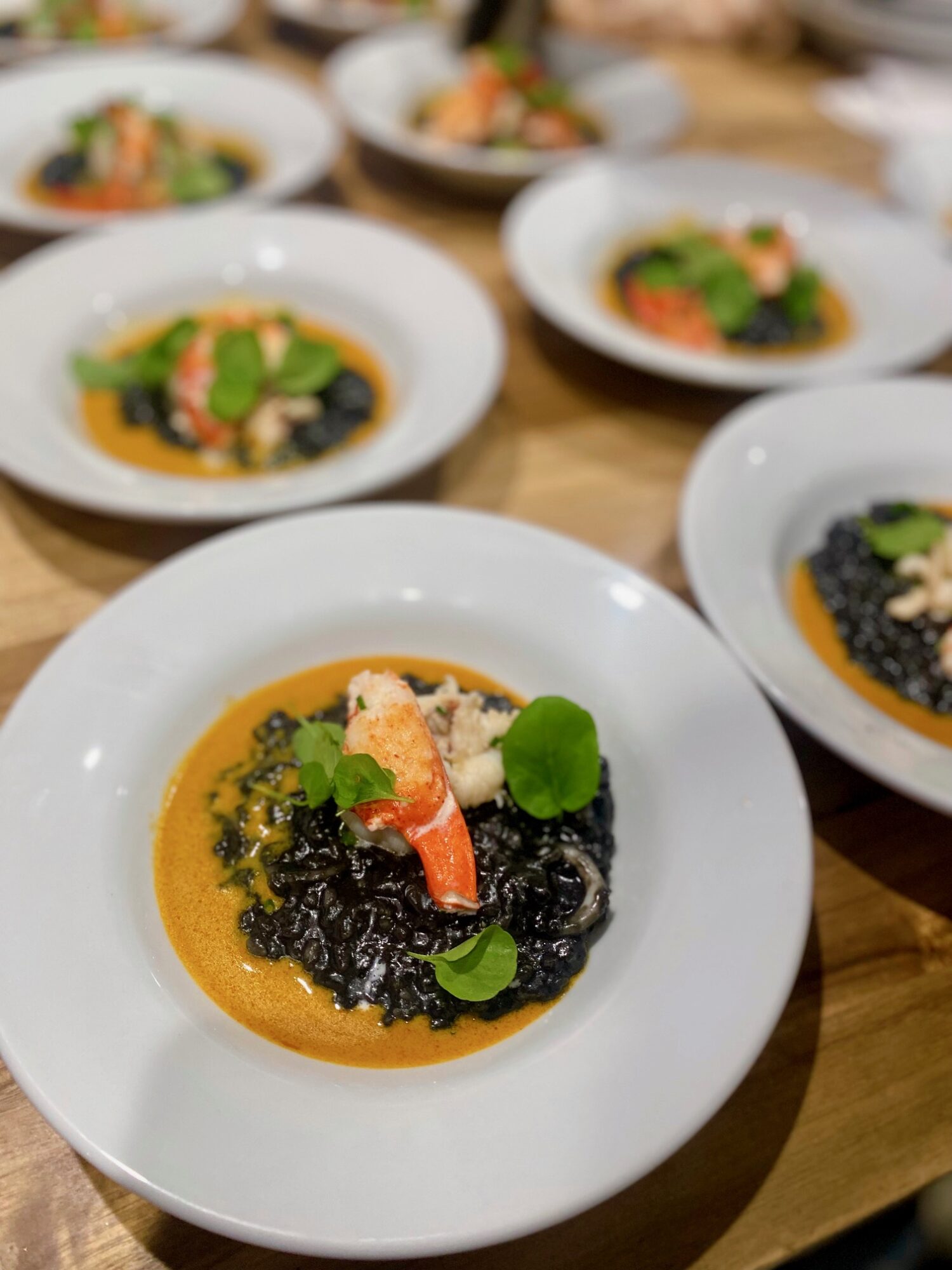 White plate with black rice, topped with shrimp, green leaves, and orange sauce, on a wooden table.