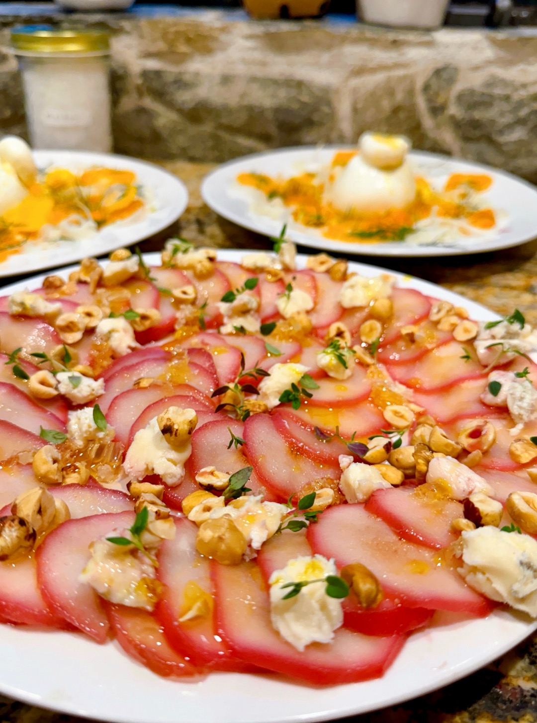 Close-up of a plate with thinly sliced cured meat, topped with cheese, herbs, and nuts, with two other plates in the background.