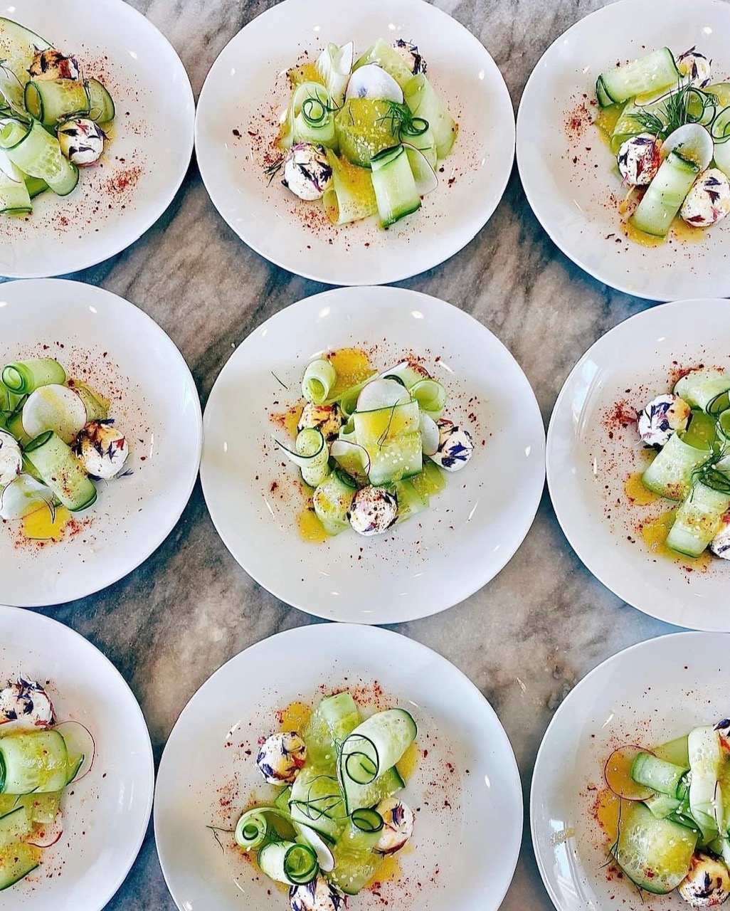 Multiple white plates with cucumber slices, small dollops of cream, and garnishes on a wooden surface.