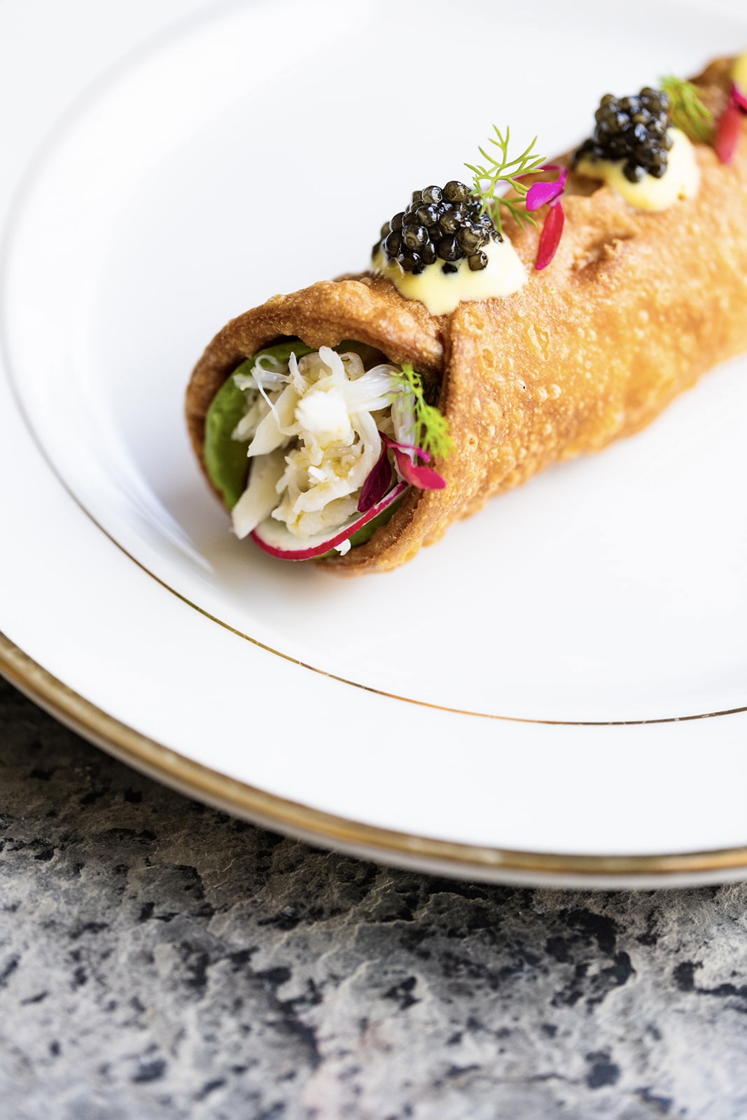 A plated appetizer with a crispy roll filled with vegetables, topped with cream, caviar, and pink garnish, on a white plate.
