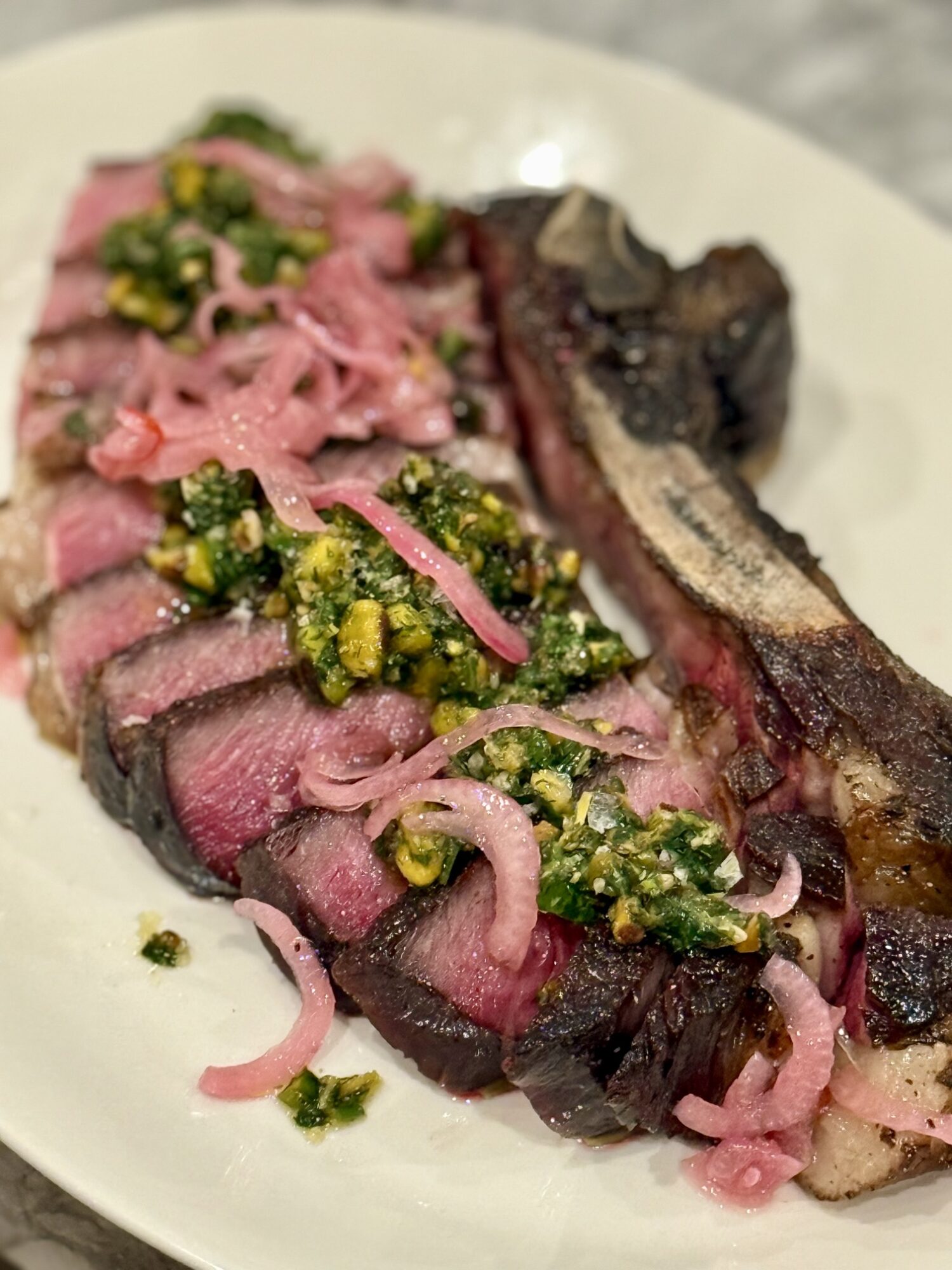 Sliced cooked steak topped with chopped herbs and pickled onions, served with a grilled bone on a white plate.