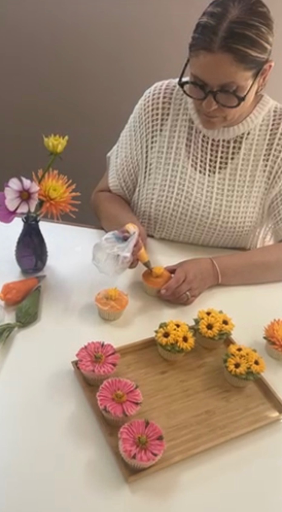 Woman arranging flowers on a table with a wooden tray and a small vase, wearing glasses and a white knit top.
