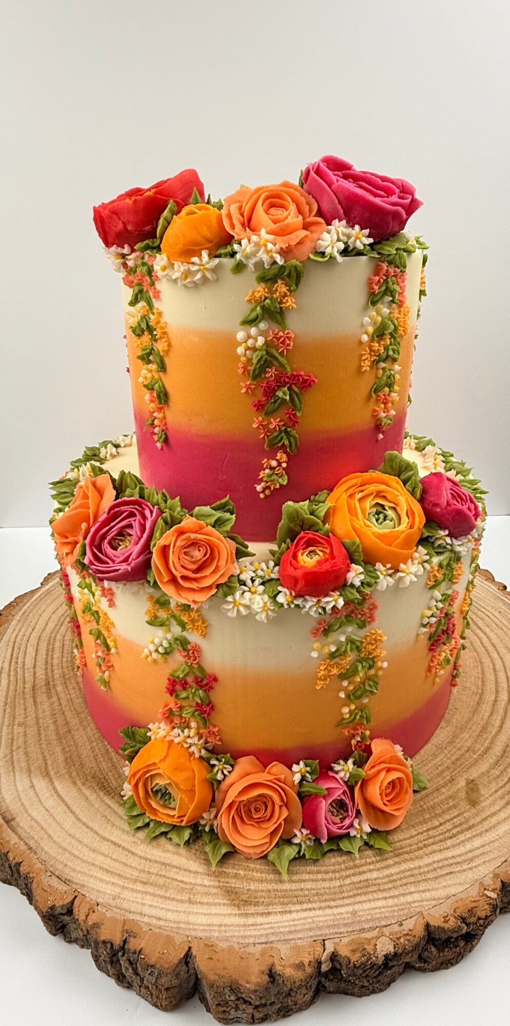 Two-tiered cake decorated with colorful roses and hanging floral garlands, placed on a wooden surface.