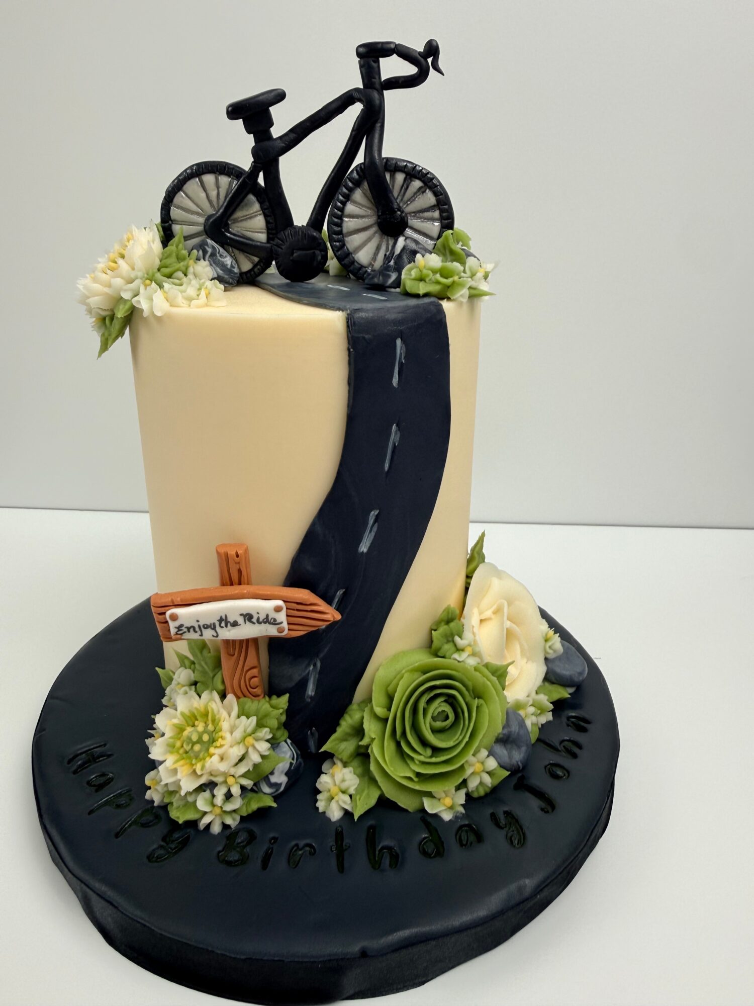 Cake with a black bicycle topper, cream and green flowers, and a black base with birthday message, on a black round board.