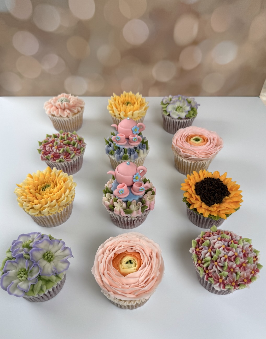 Twelve decorated cupcakes arranged in three rows on a white surface, with a blurred background. Each cupcake has colorful frosting and decorations.