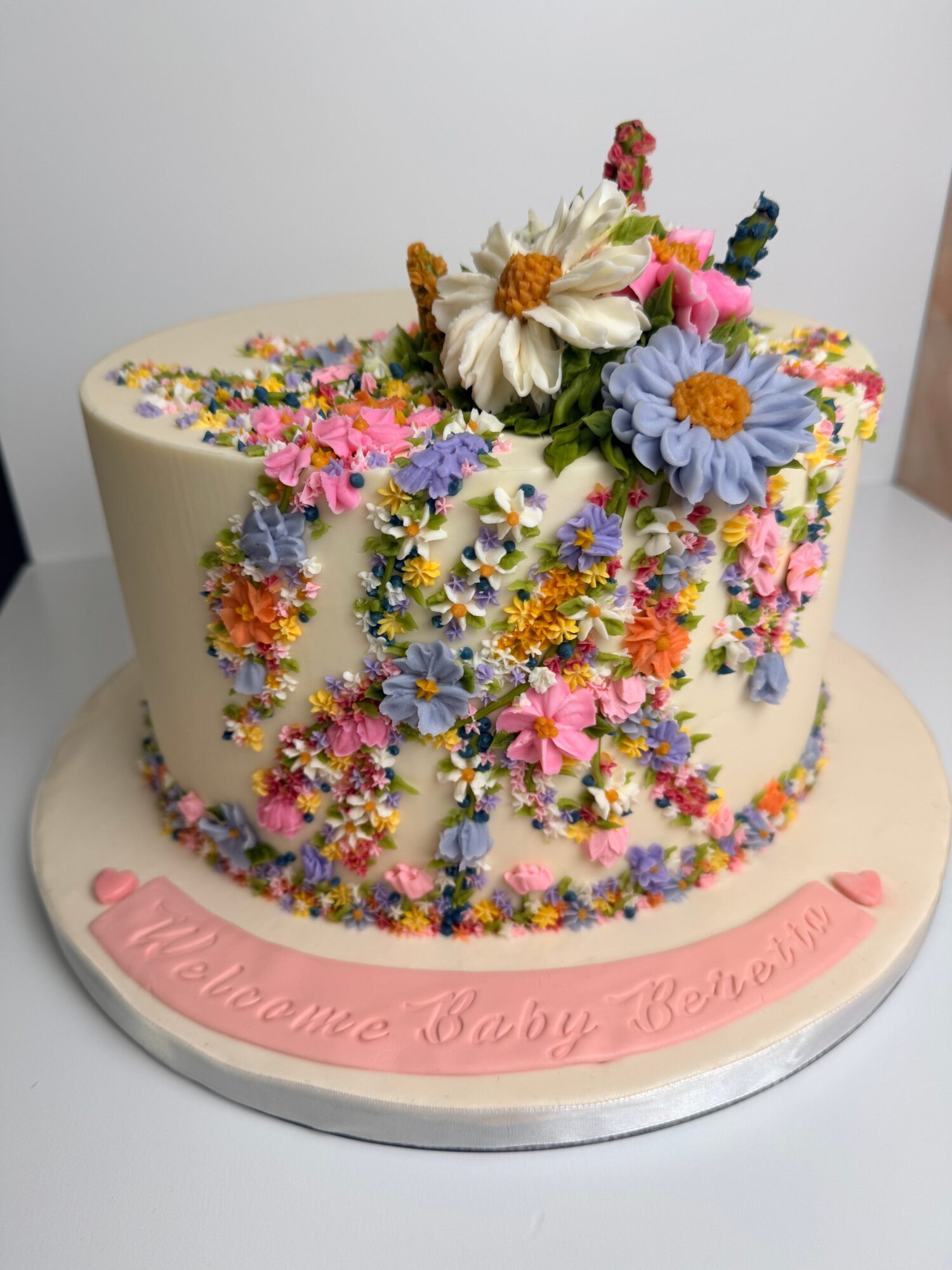 Round cake decorated with colorful flowers and a pink ribbon with text, on a white surface.